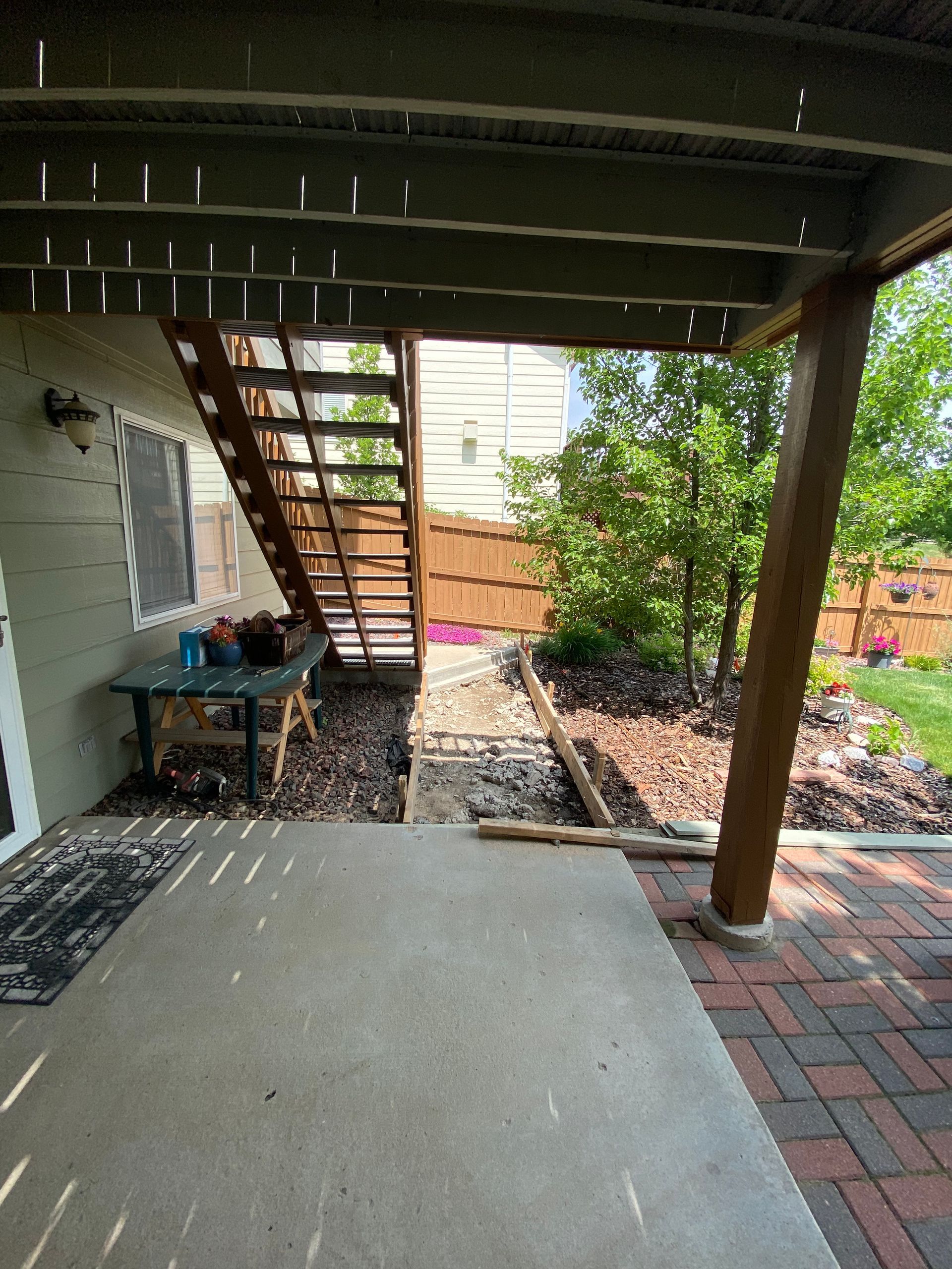 Concrete patio leading to stairs under a deck and a brick pathway surrounded by landscaping.