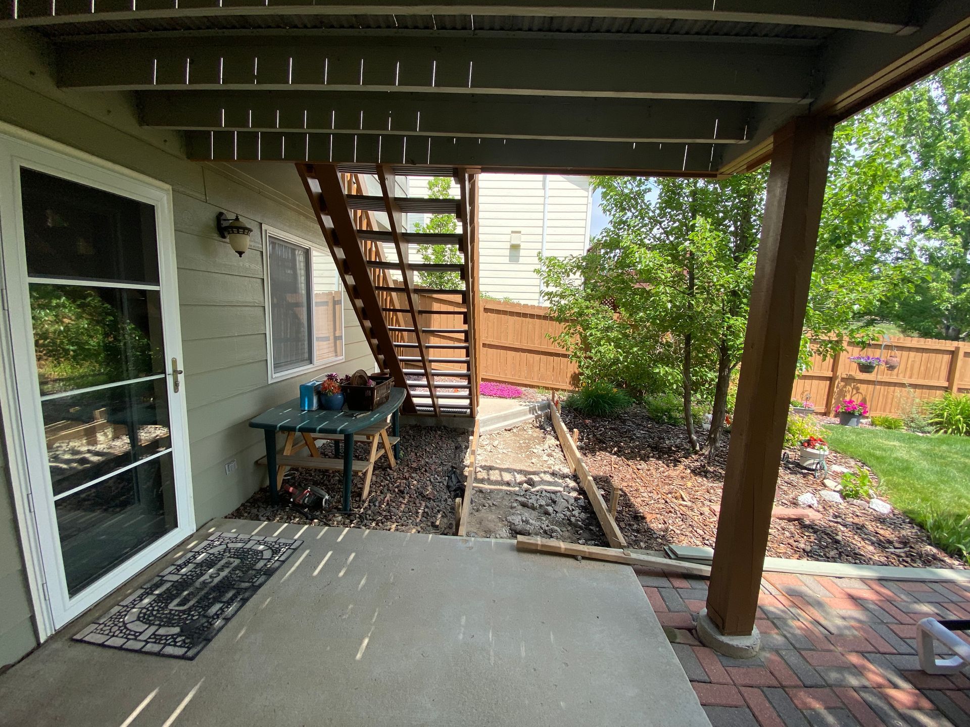 Backyard patio with stairs leading up, a door, a table, and a brown fence.