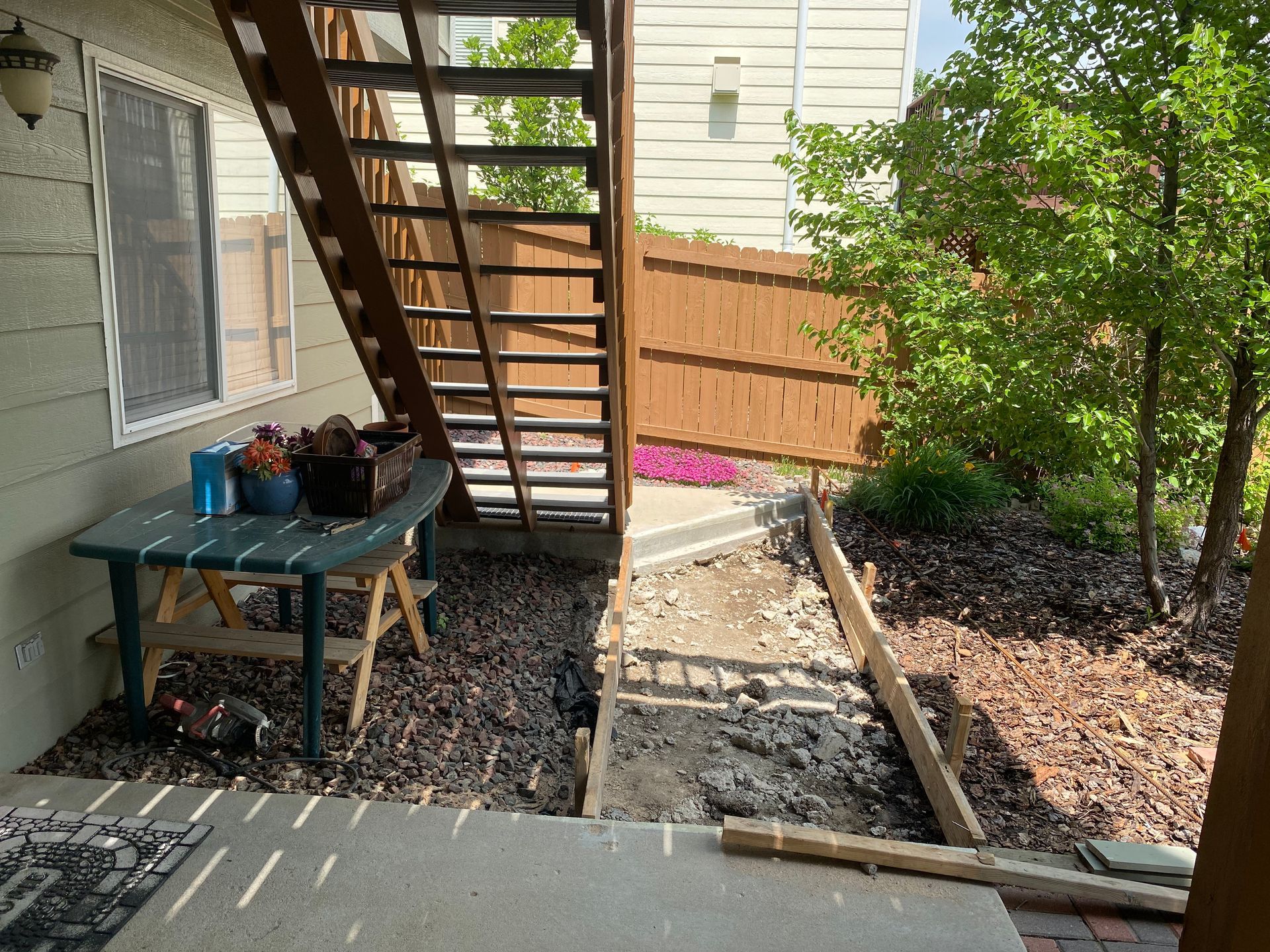 Backyard with stairs, table with items, construction underway along a pathway, surrounded by mulch, fence, and a tree.