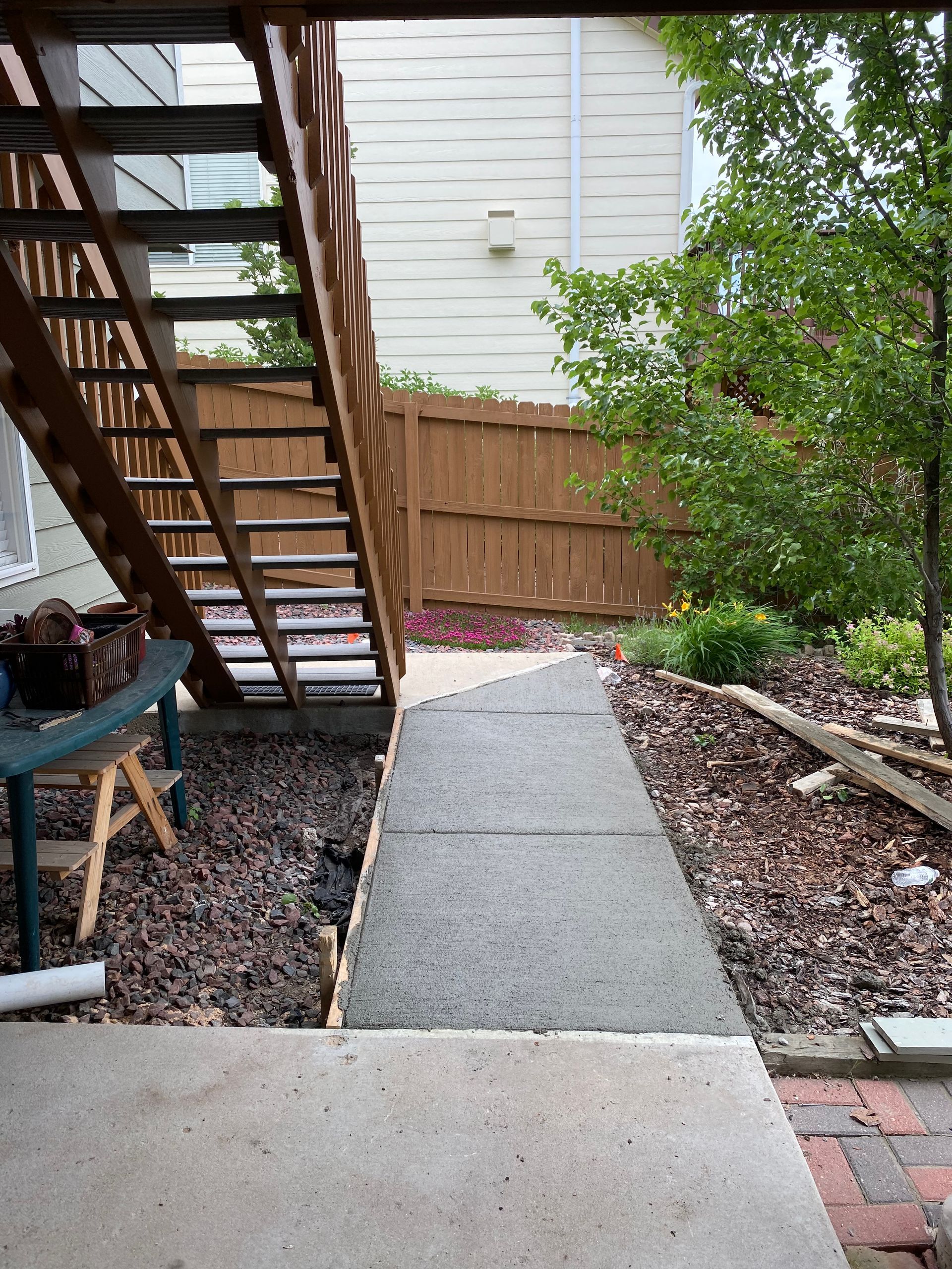 Concrete walkway leading under wooden stairs, through gravel, to a fenced yard.