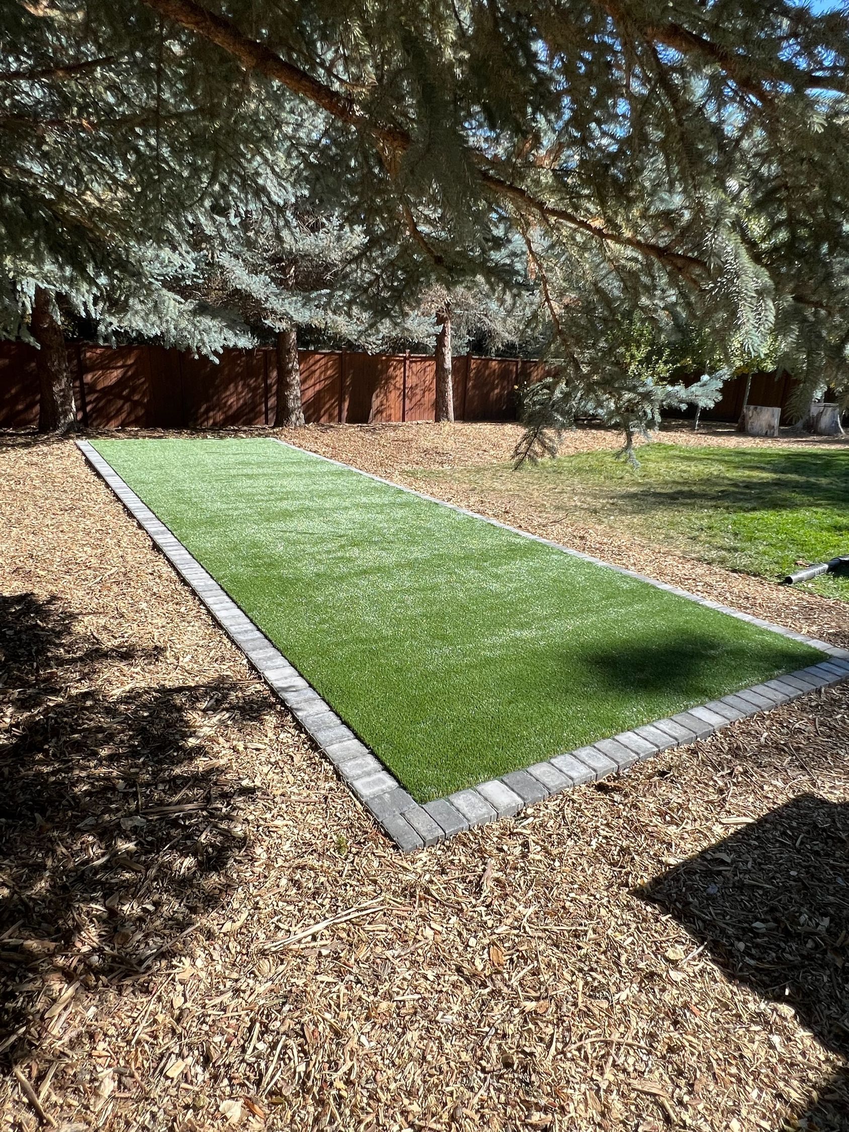 A rectangular patch of artificial grass bordered by pavers, surrounded by wood chips, under trees.