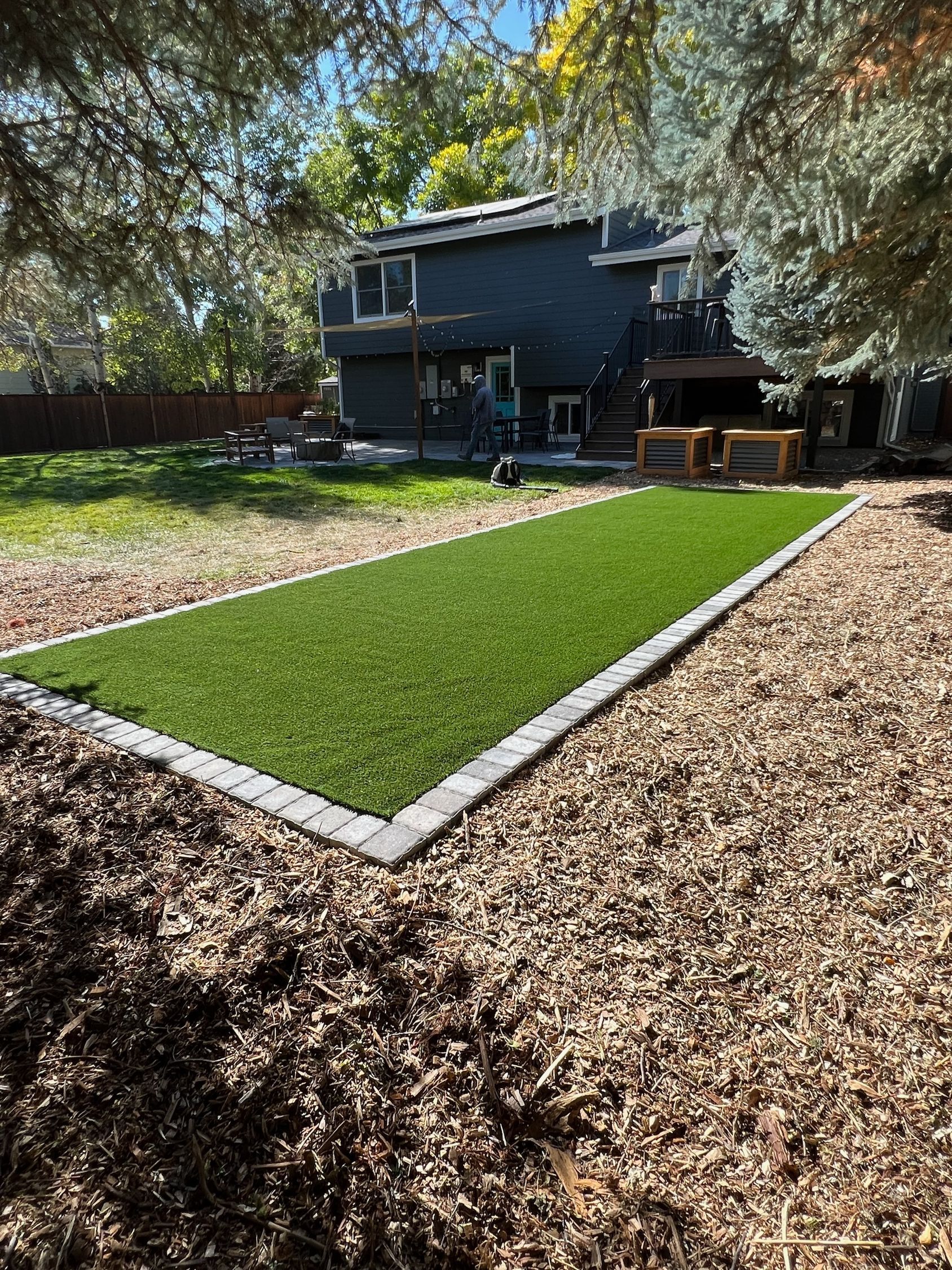 Backyard with a rectangle of green turf bordered by pavers; surrounded by wood chips, trees, and a house.