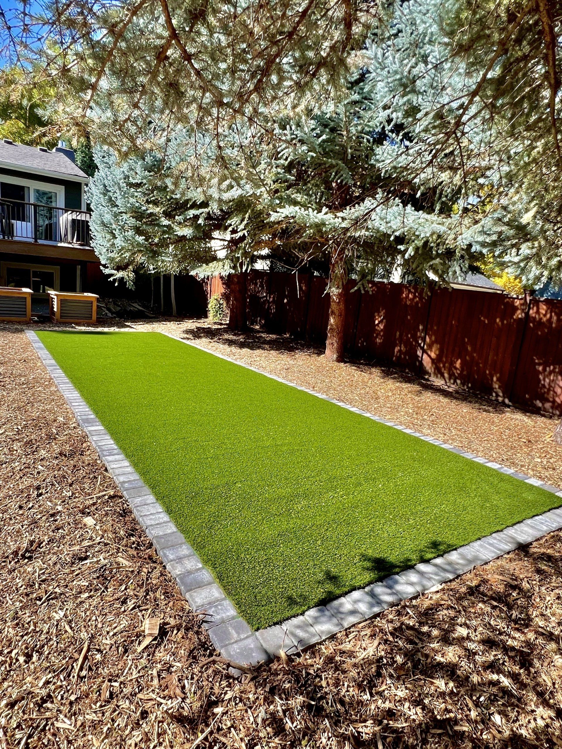 Backyard with rectangular artificial turf, bordered by paving stones. Brown mulch, trees, and a wooden fence surround it.