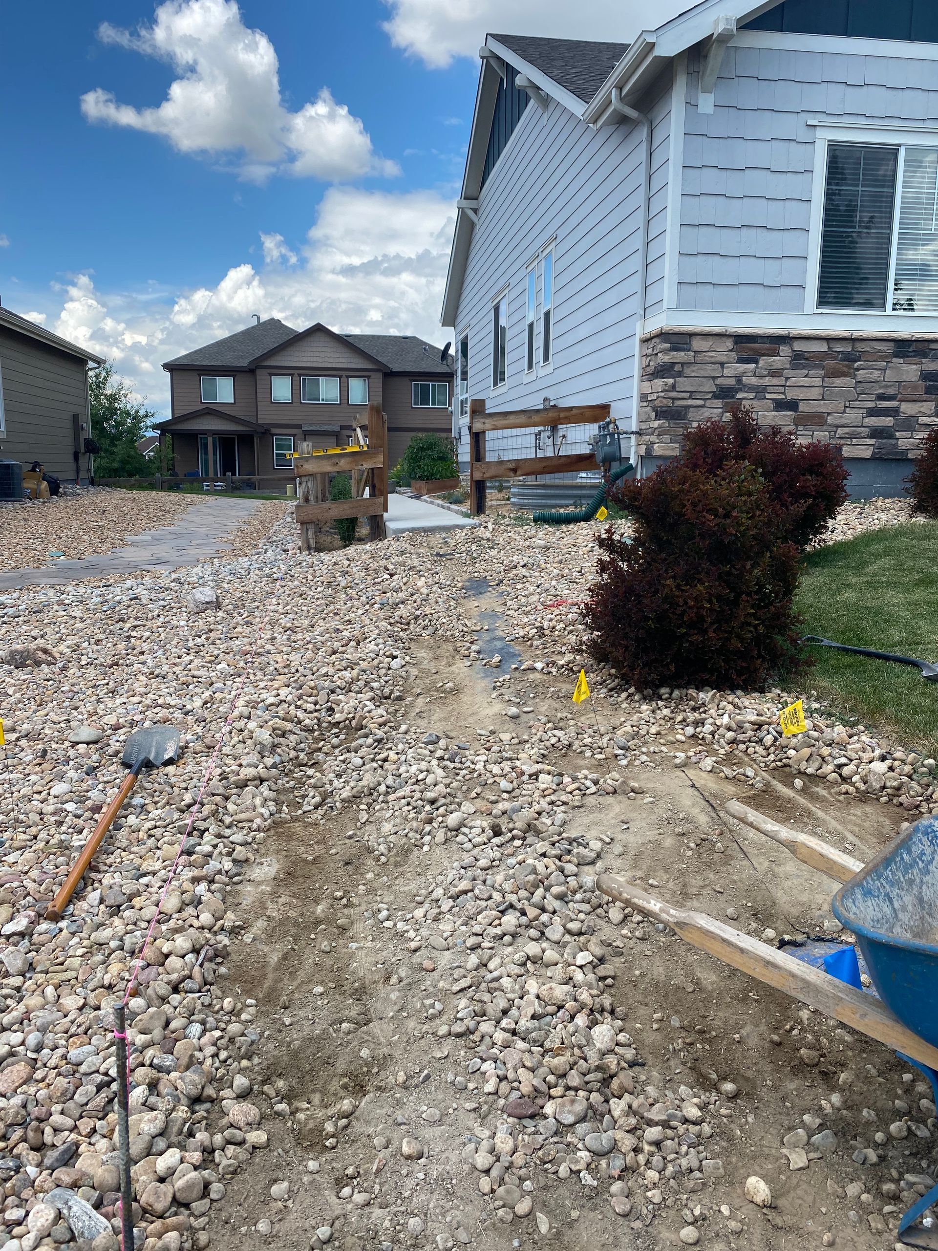 Construction site with gravel pathway leading to a house, with wooden barriers and a wheelbarrow. Blue sky overhead.