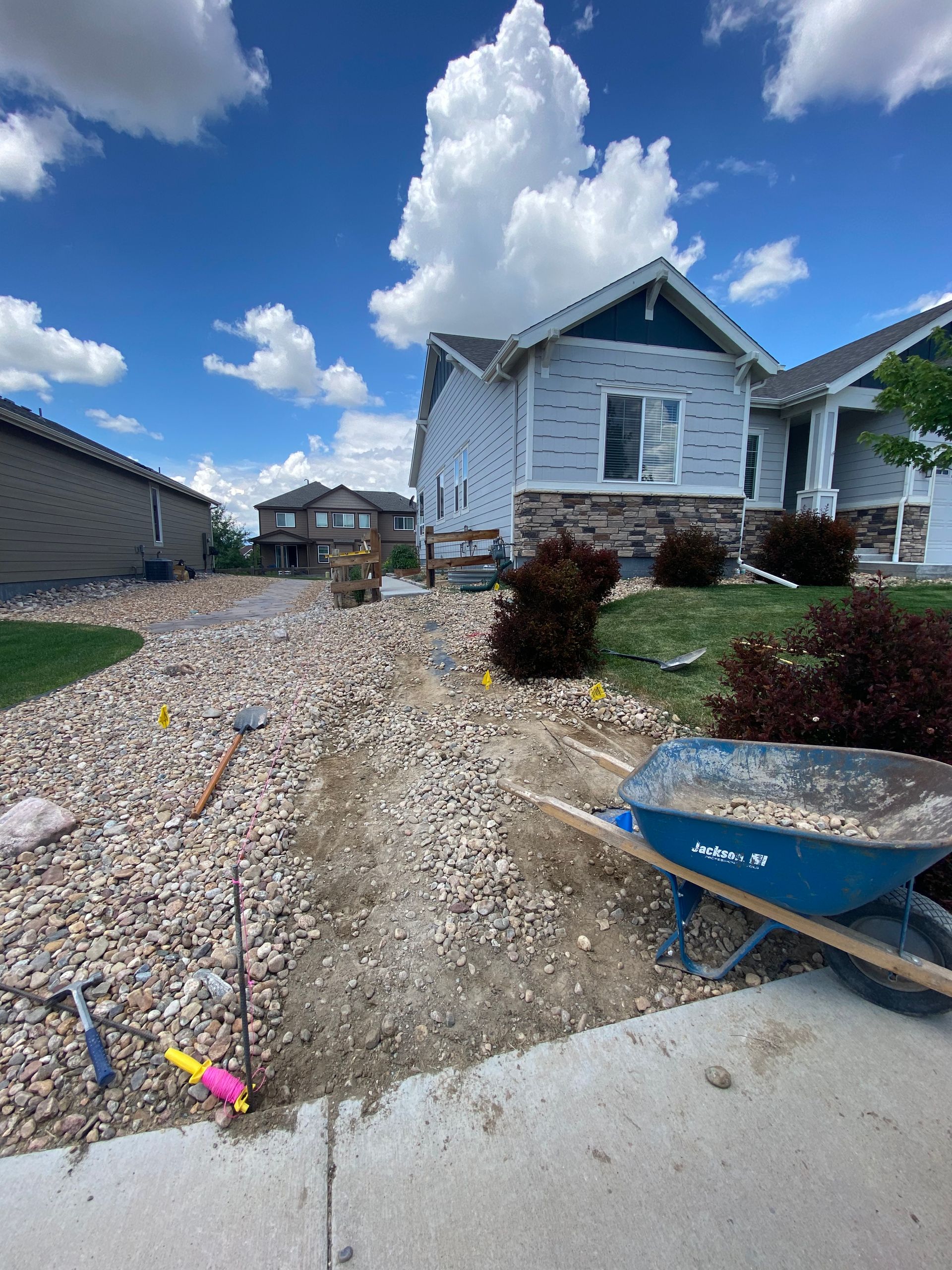 Construction of a rock pathway in front of a blue house with a wheelbarrow and tools present on a sunny day.