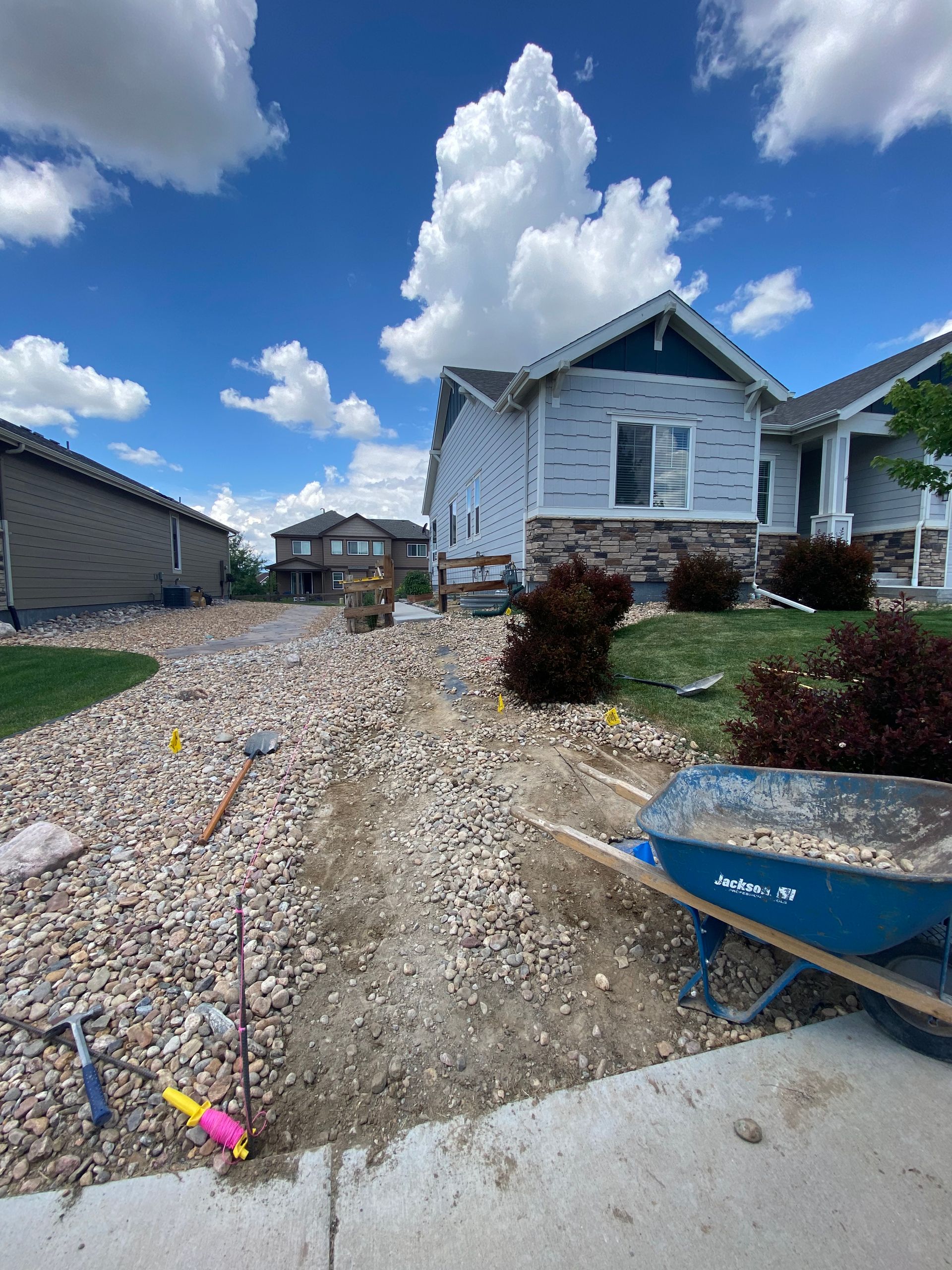 Gravel pathway under construction at a house with blue siding. A wheelbarrow and tools are visible. Sunny day.