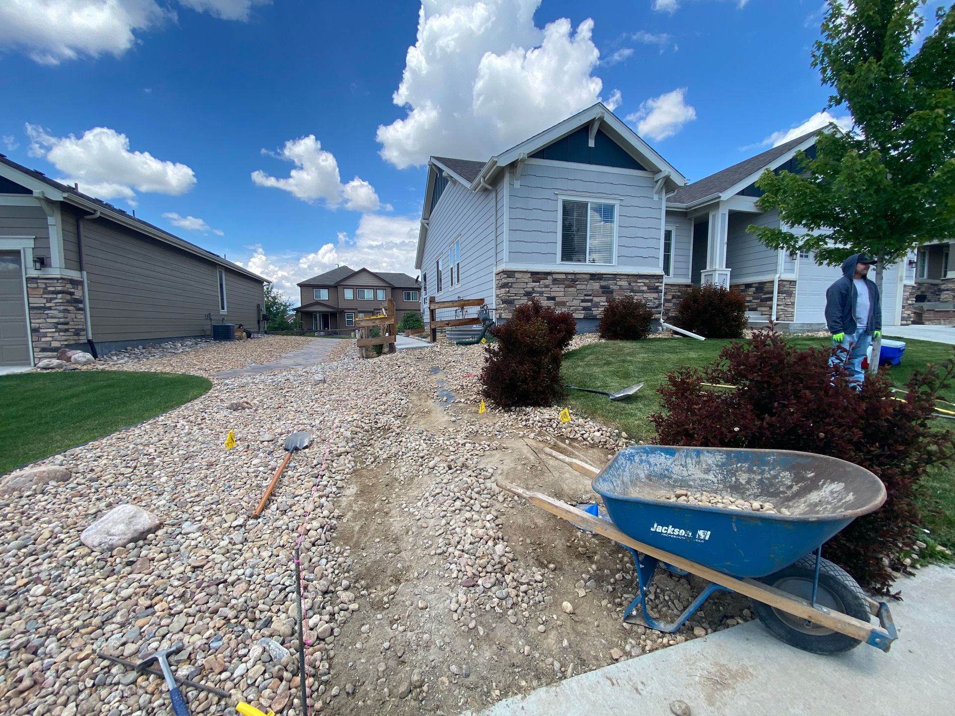A house with gravel landscaping; a wheelbarrow, tools, and a worker are present.