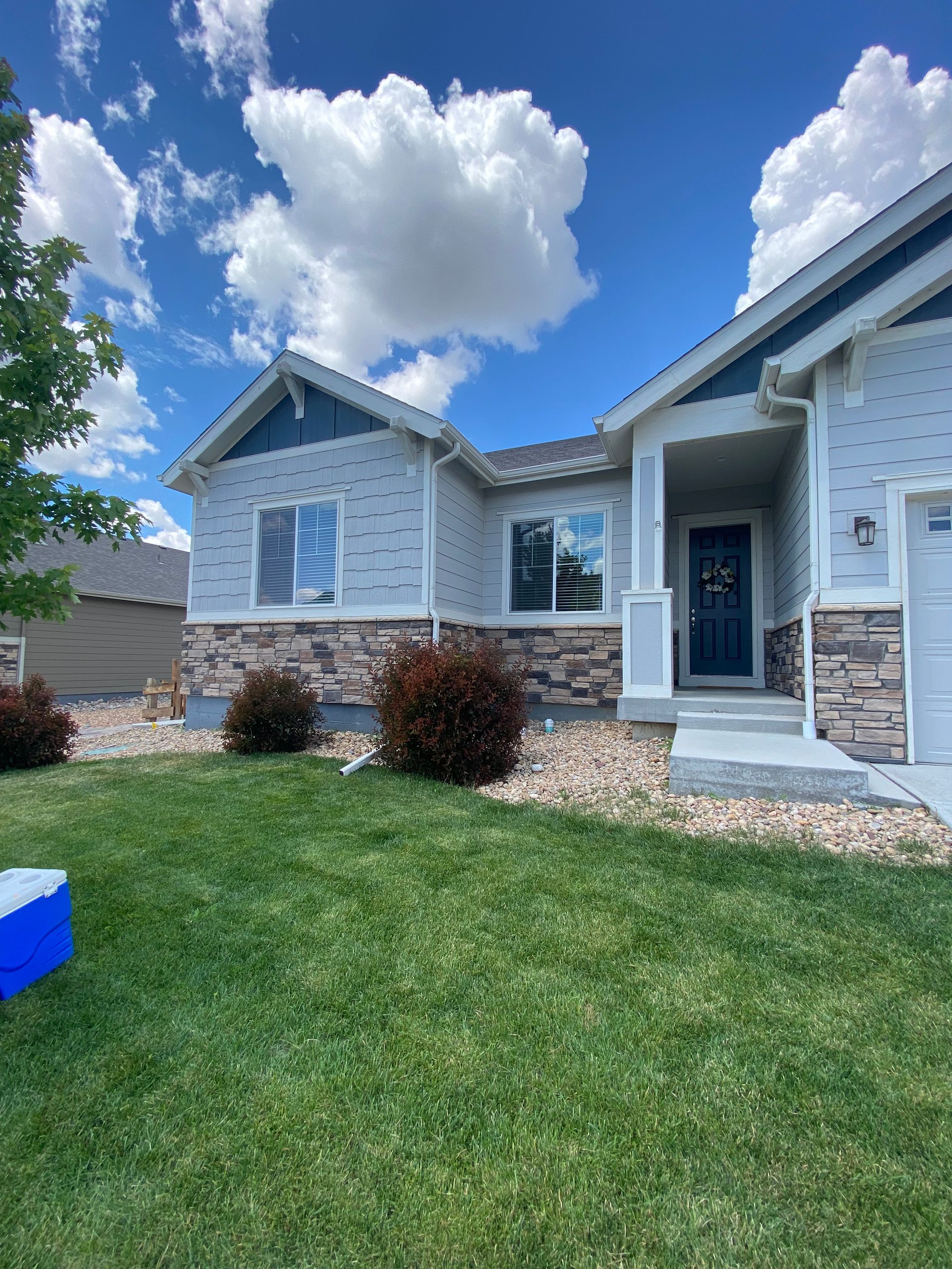Light blue house with stone accents, blue trim, and lush green lawn under a partly cloudy sky.