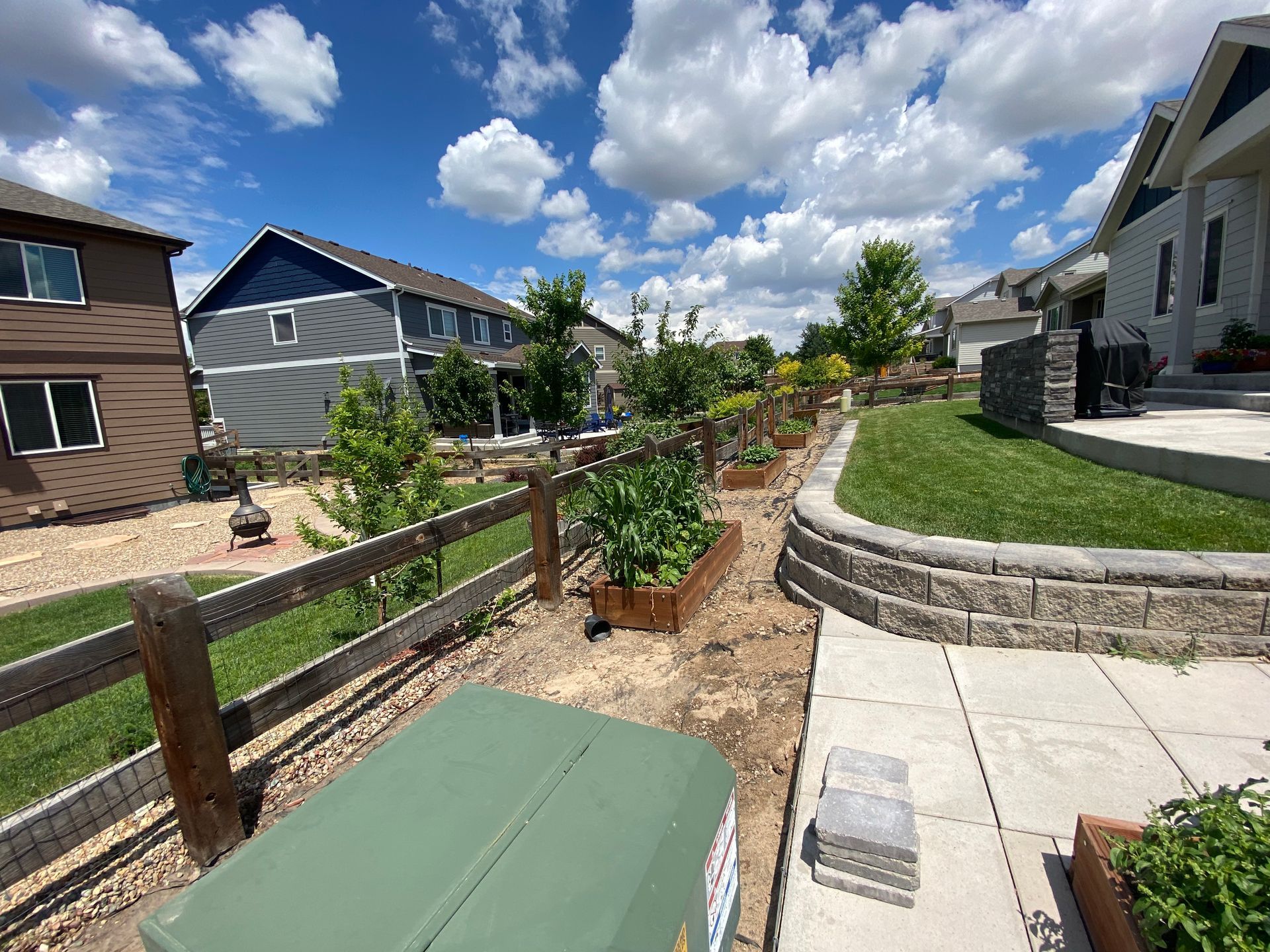 Backyard garden with raised beds and wooden fence, under blue sky with clouds.