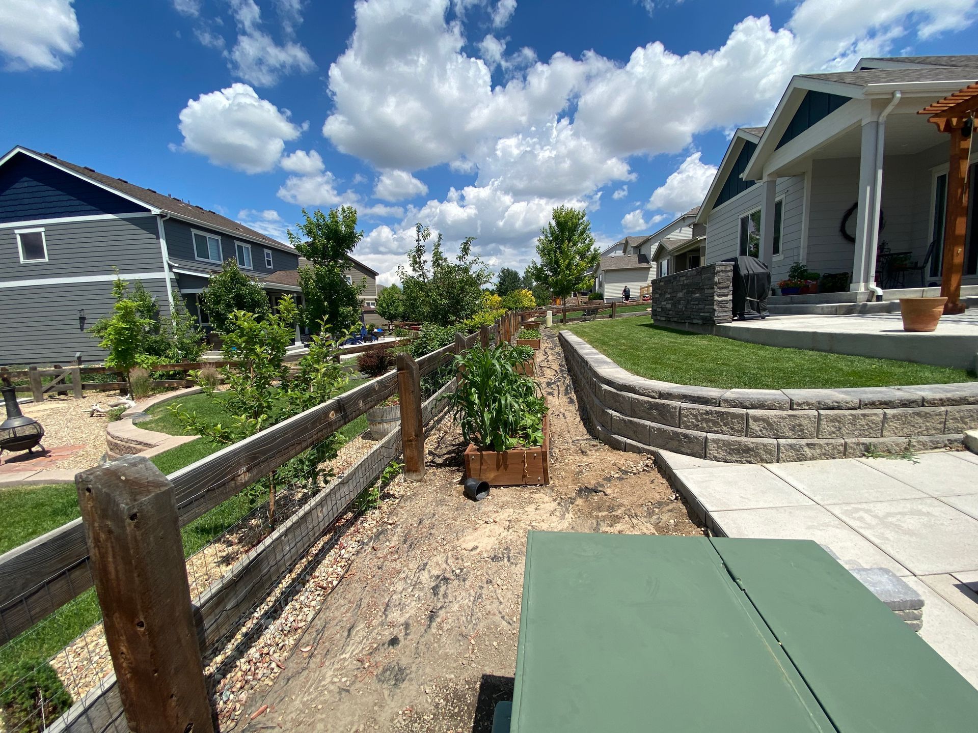 Backyard with garden beds, a fence, houses, and a picnic table on a sunny day.