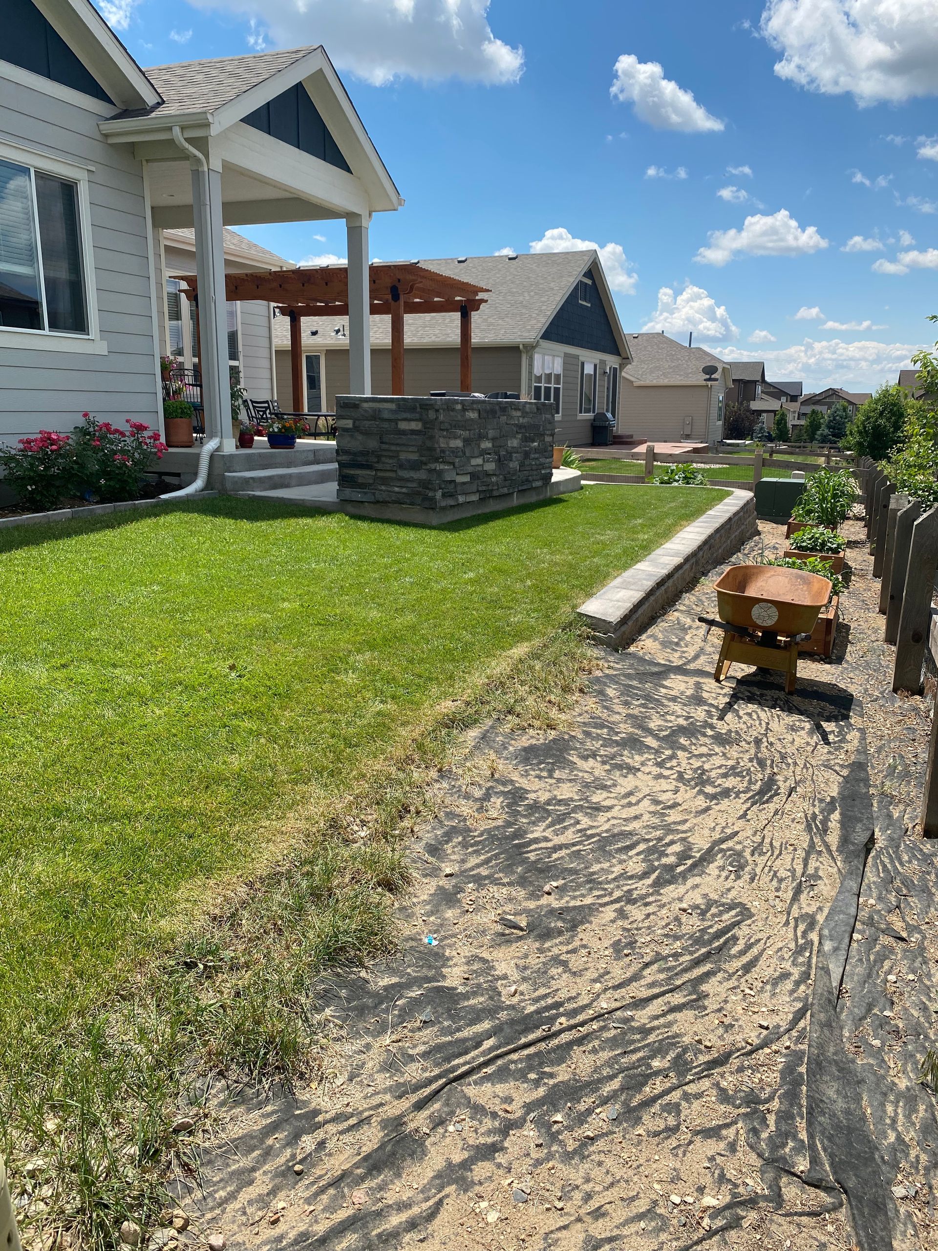Lawn and garden scene with house, stacked stone feature, retaining wall, and fire pit under blue sky.