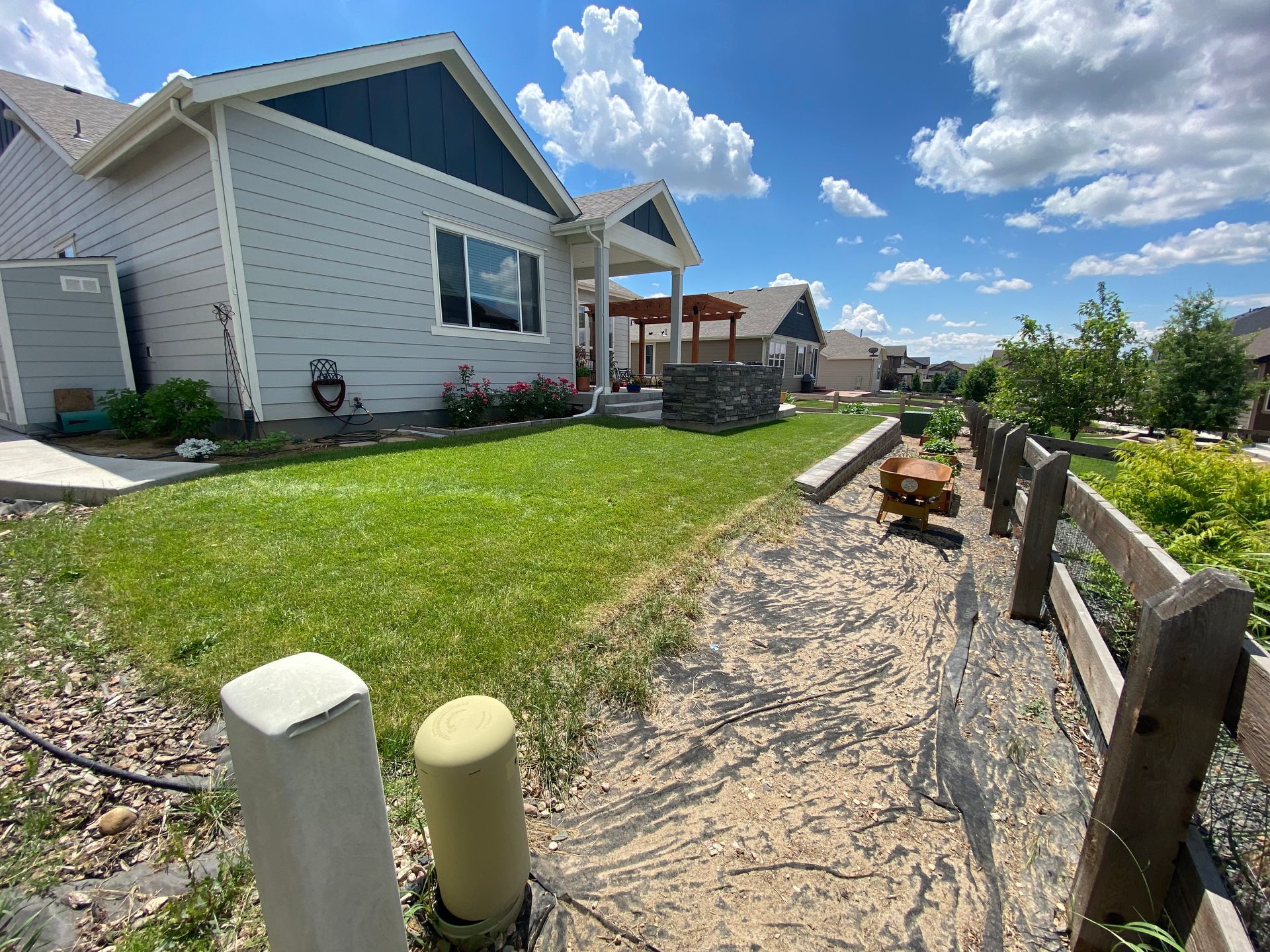 A light-colored house with blue trim and a green lawn on a sunny day. A wooden fence runs along a sandy path.
