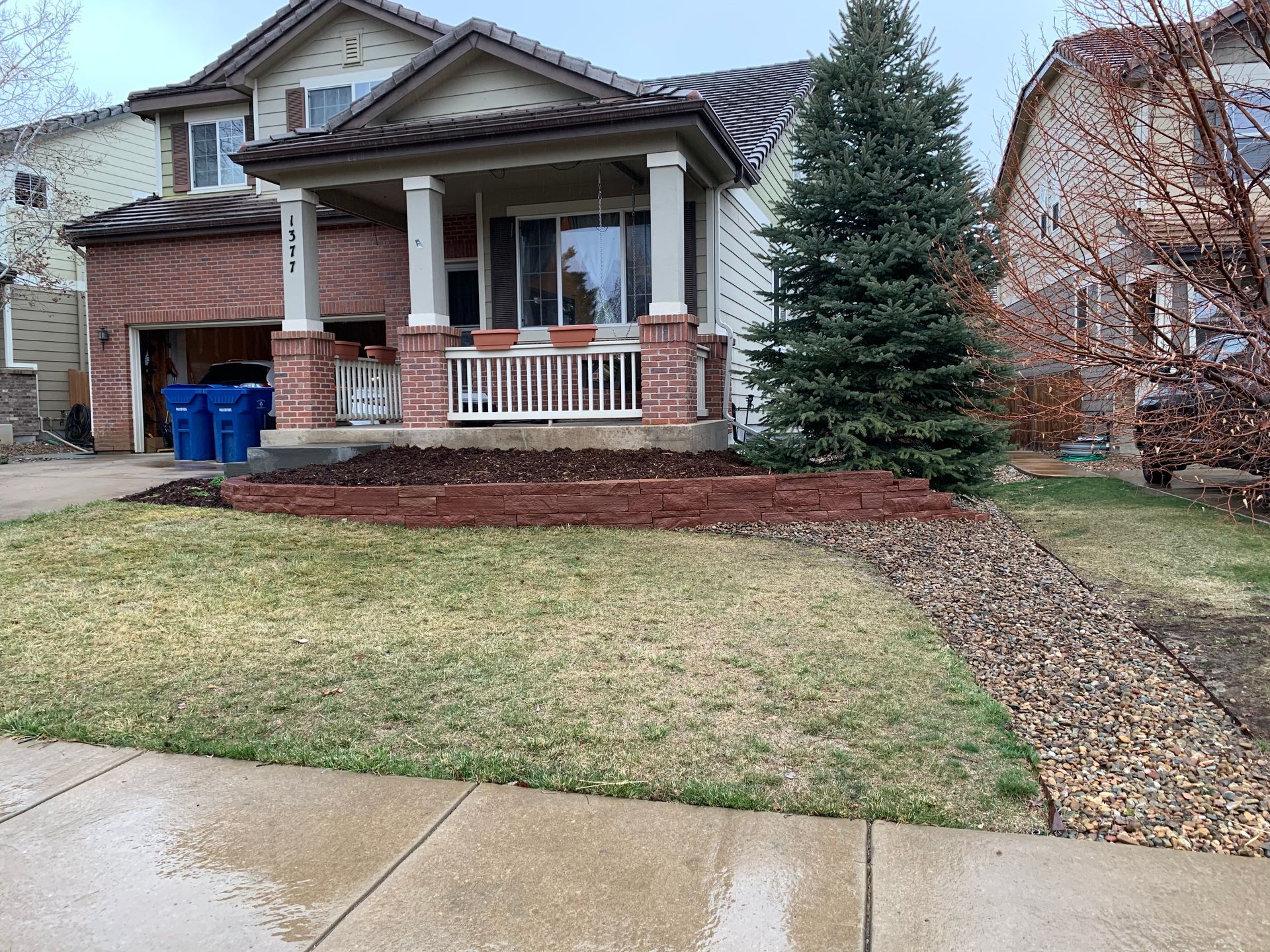 A two-story house with a small porch and an evergreen tree. Brown brick garden bed, patchy grass.