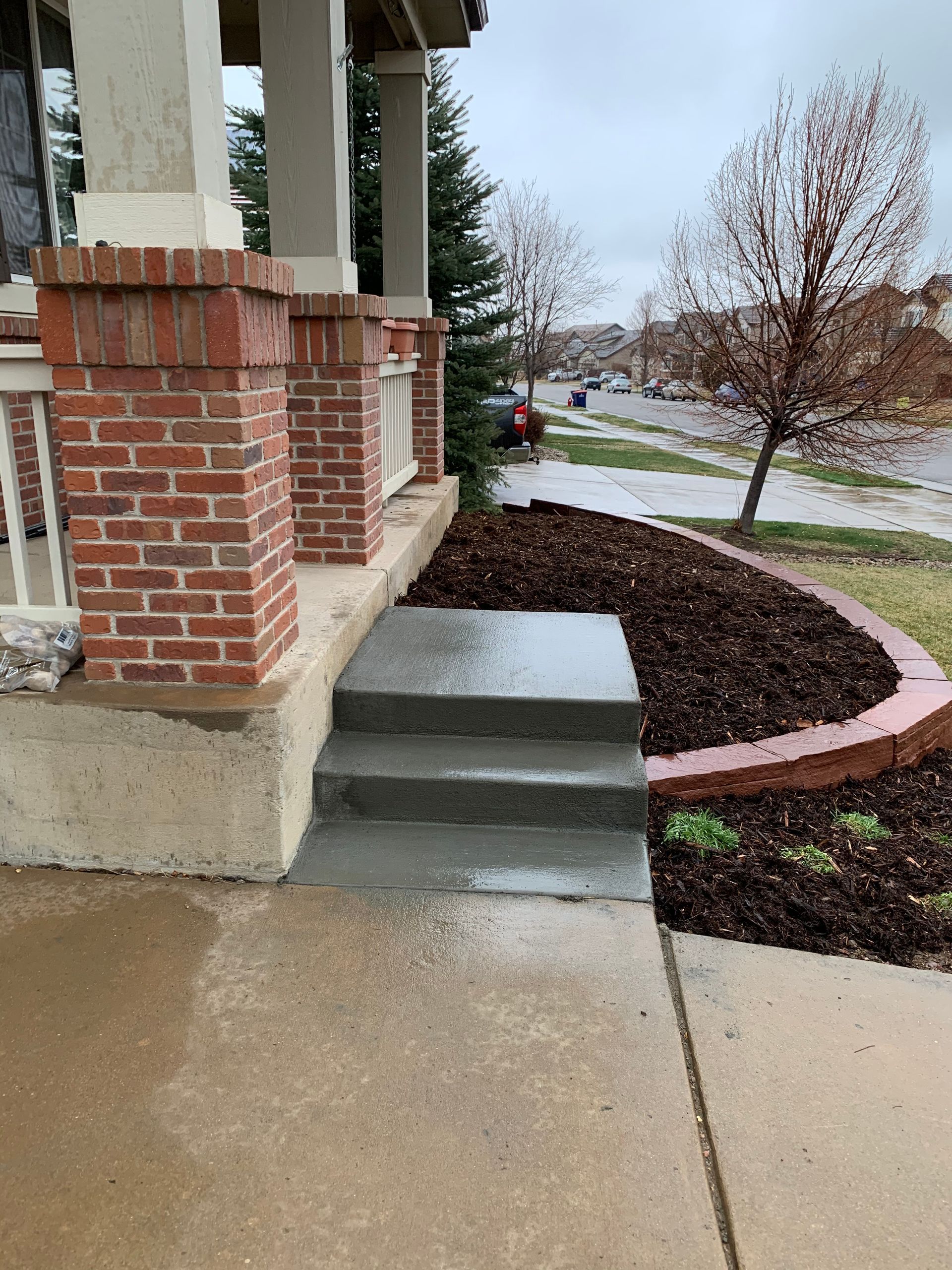 Concrete steps leading to a brick-columned porch with a mulch-covered garden bed, a cloudy day.