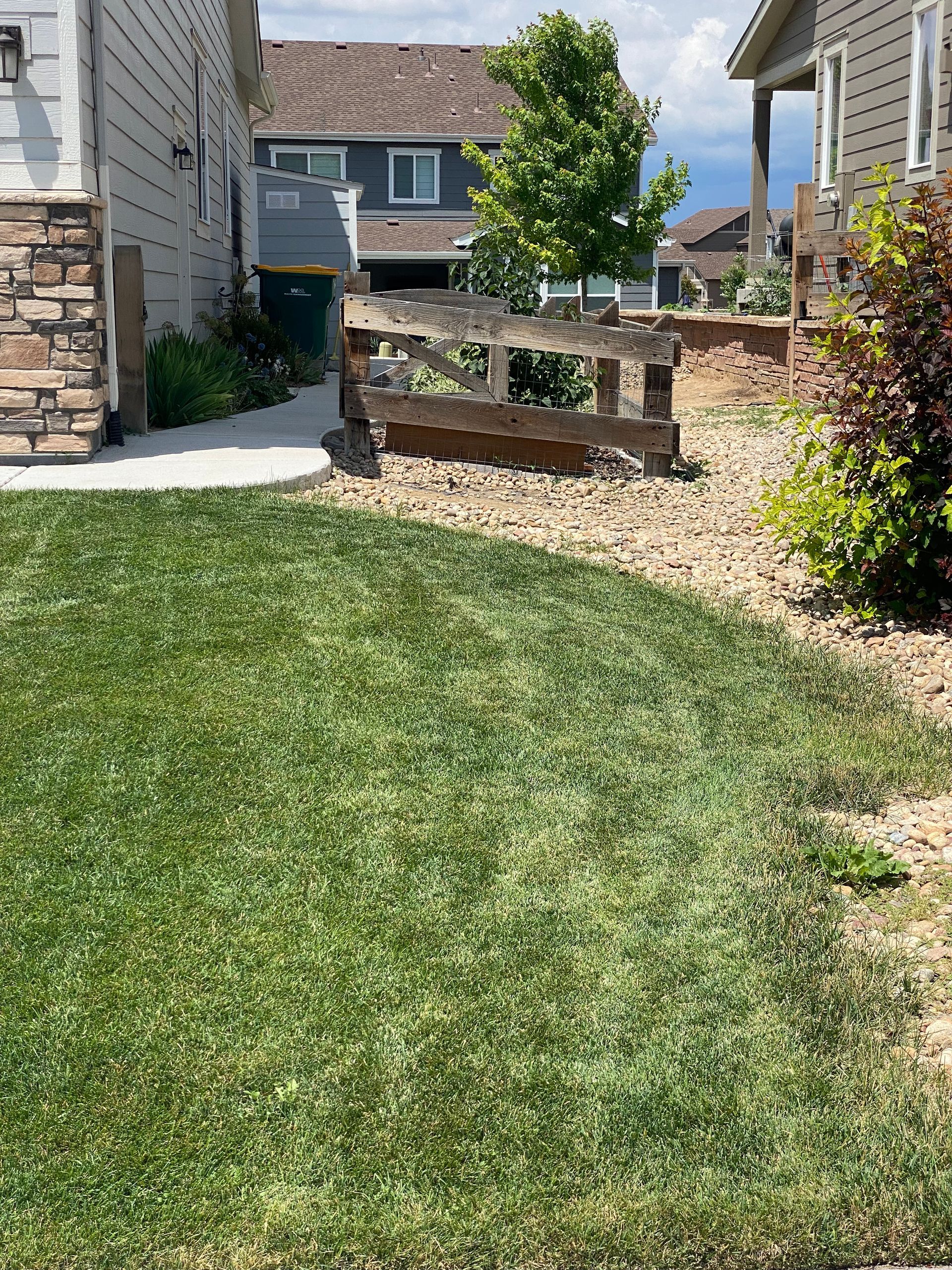 Lush green lawn in front of a house. A weathered wooden fence separates the grass from a rock bed and a neighboring house.