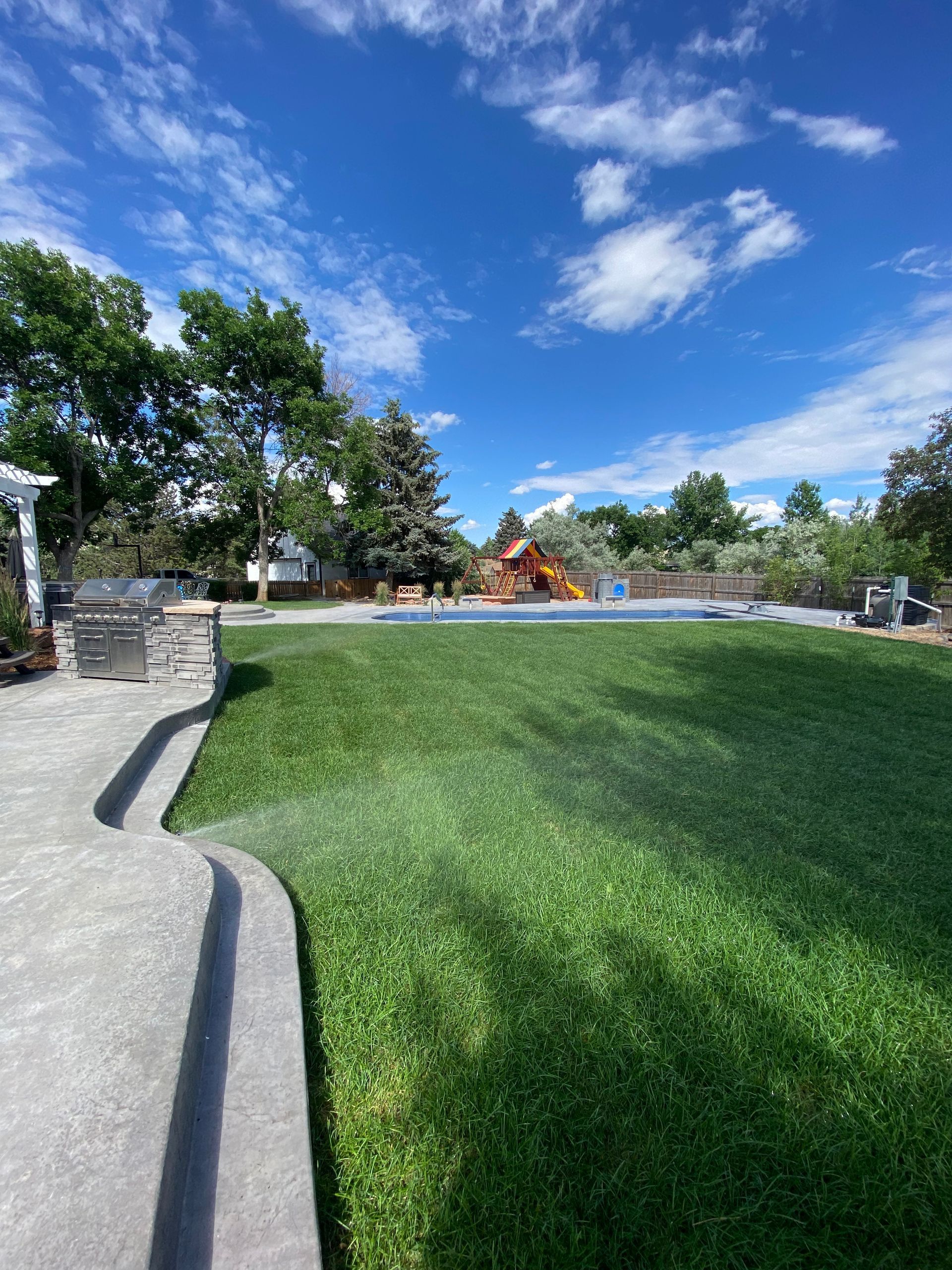 Lush green lawn with sprinkler watering, bright blue sky with clouds, and a pool in the distance.