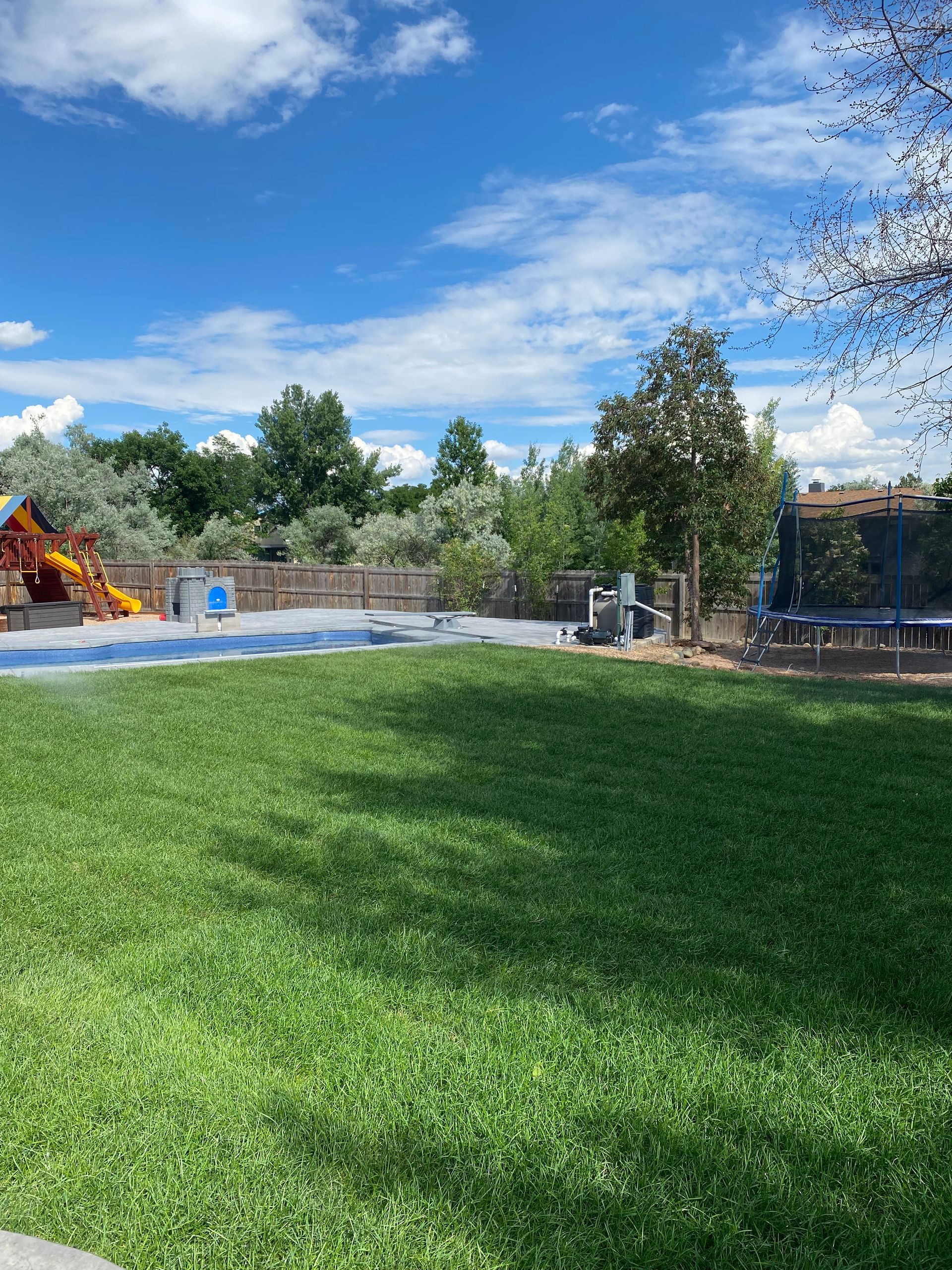 Grassy lawn with a partially visible pool and playground under a blue sky with fluffy clouds.