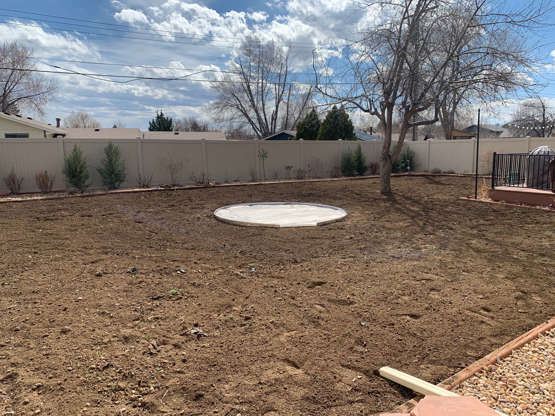 Backyard with dirt ground, a concrete patio, trees, and fence on a sunny day.