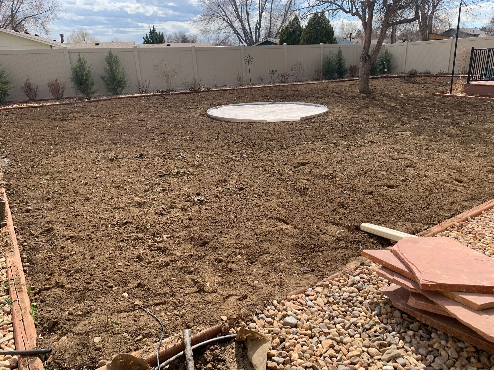 Backyard with prepared soil, circular concrete pad, and white fence.