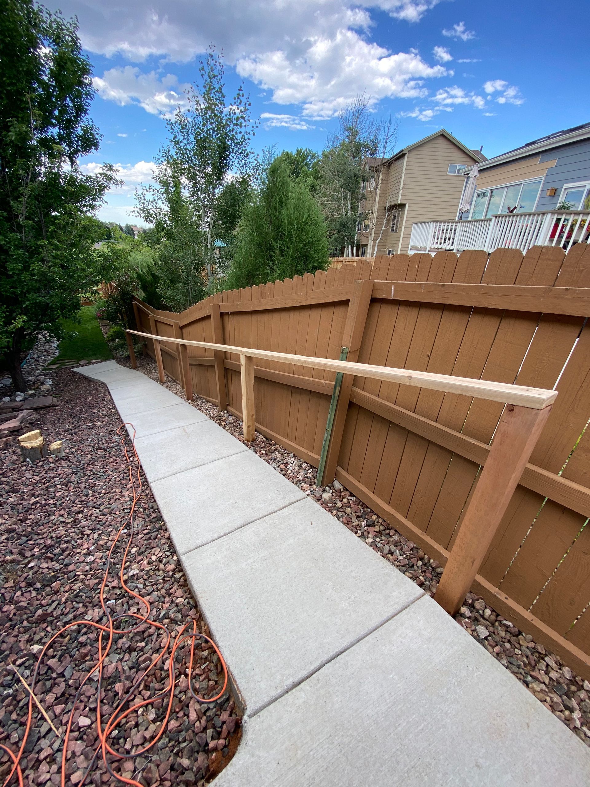 Concrete path alongside a wooden fence, with a handrail. Trees and a house are visible in the background under a blue sky.