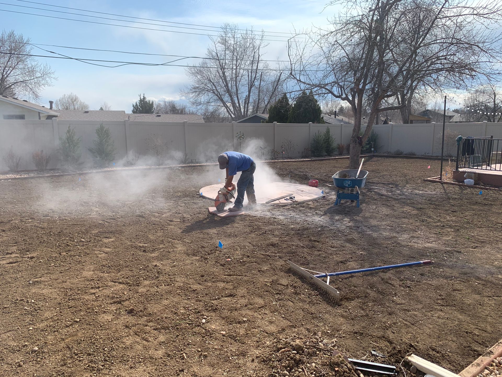 Man cutting concrete in a backyard, creating dust. The yard is mostly dirt, with some tools visible.