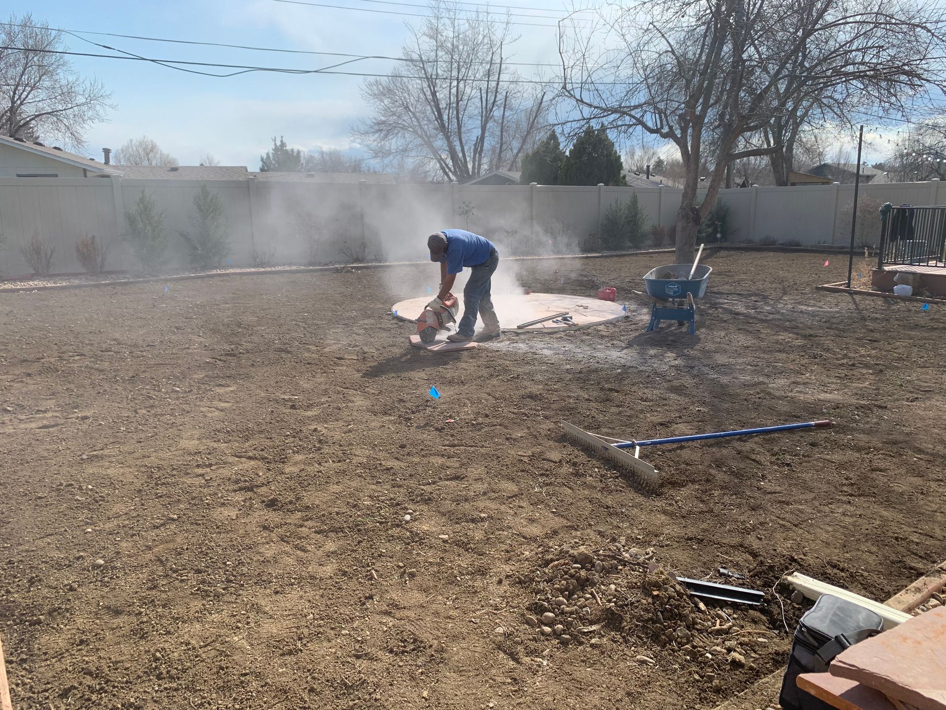 A person cutting a concrete patio in a backyard, creating a cloud of dust.