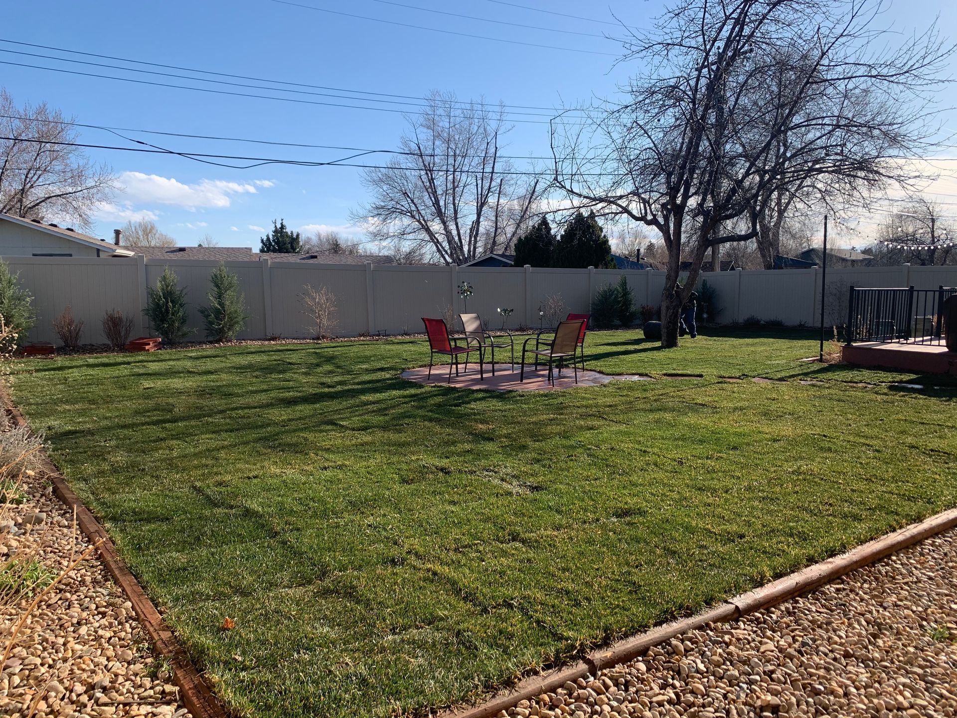 A green backyard with a fire pit, seating, and a beige fence under a blue sky.