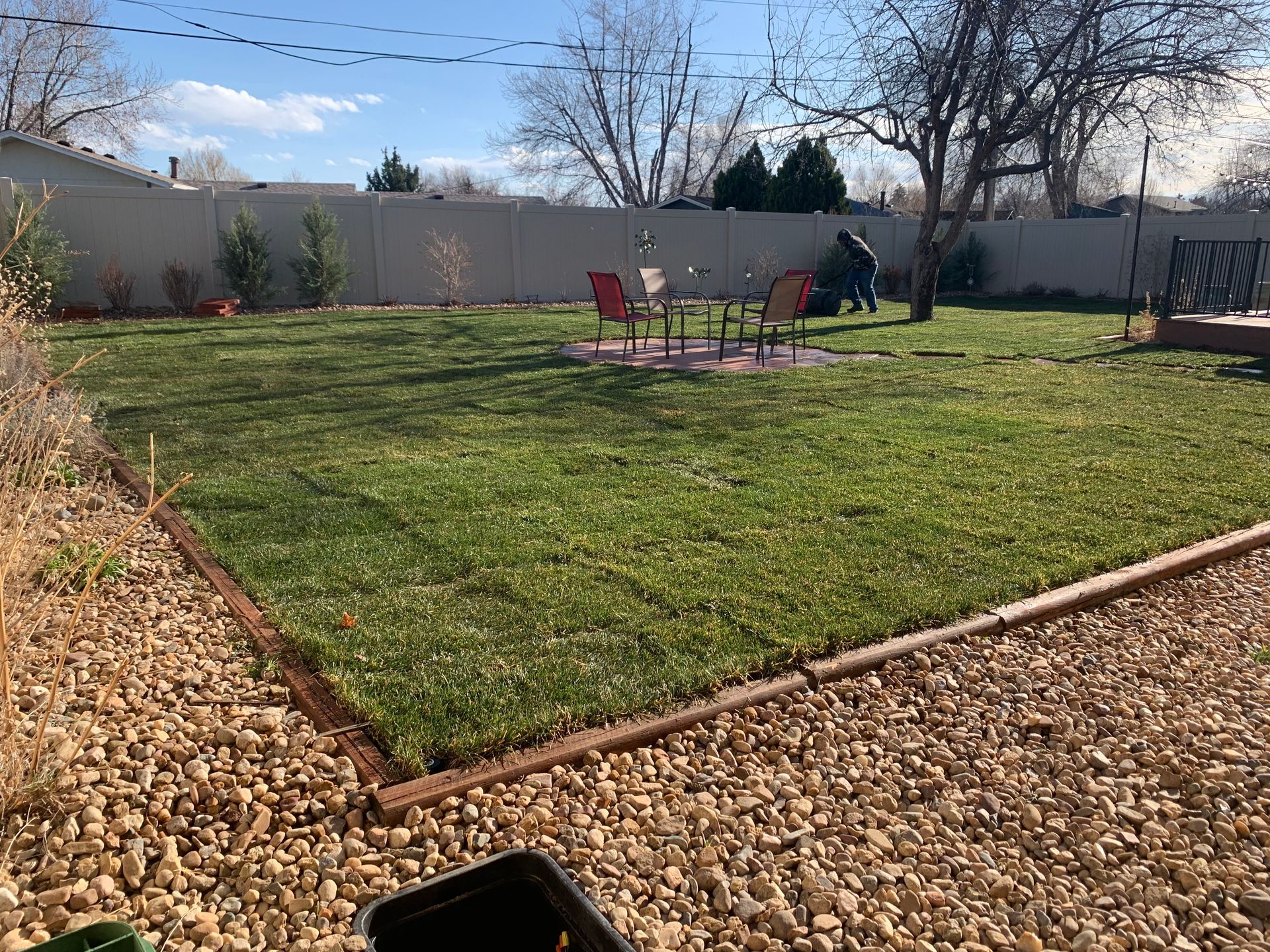 Lush green lawn with gravel border and a table and chairs, person mowing, fenced backyard.