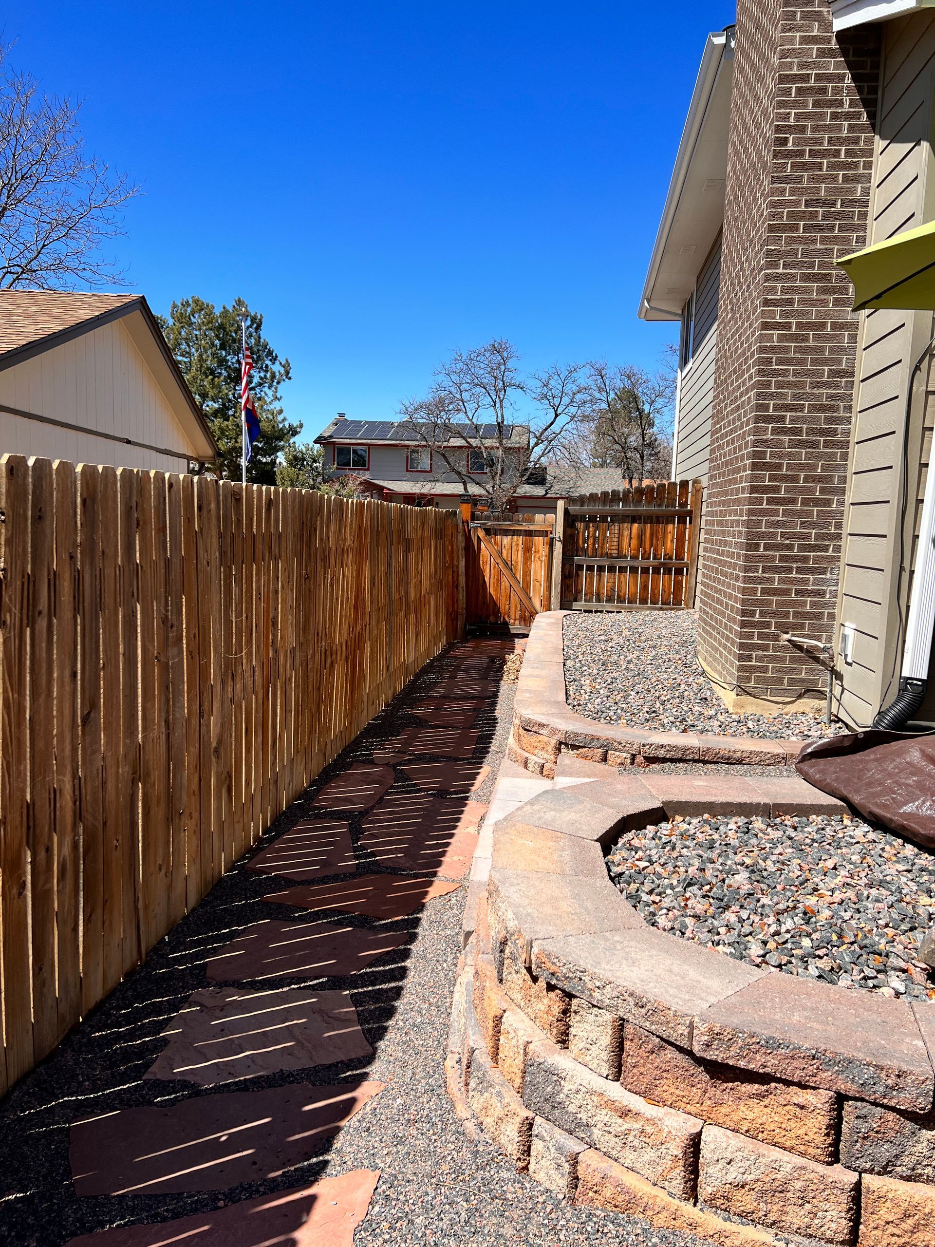 Narrow backyard pathway with stone pavers and retaining wall, fenced on one side.