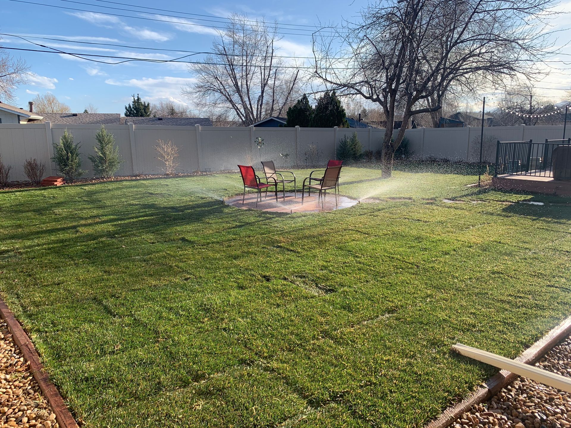 Sprinkler watering a green lawn in a backyard with chairs, a fence, and trees under a blue sky.