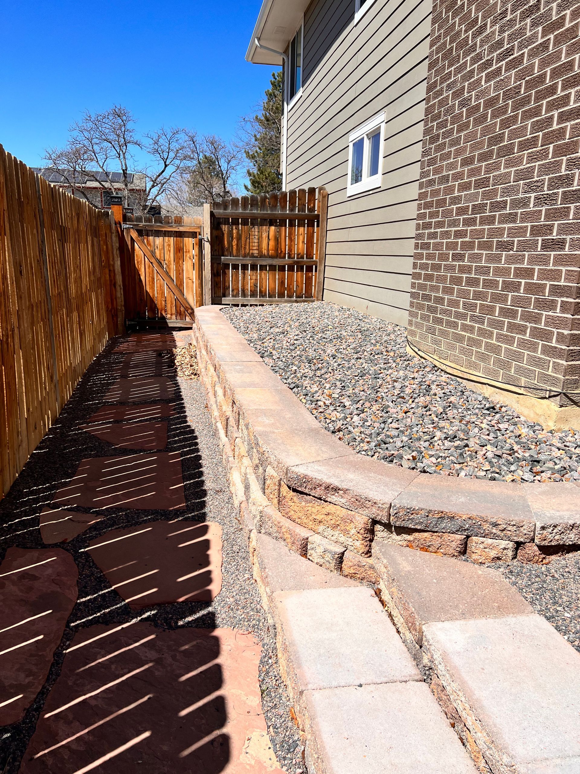 Pathway beside a house, between a wooden fence and a brick wall with rock landscaping.