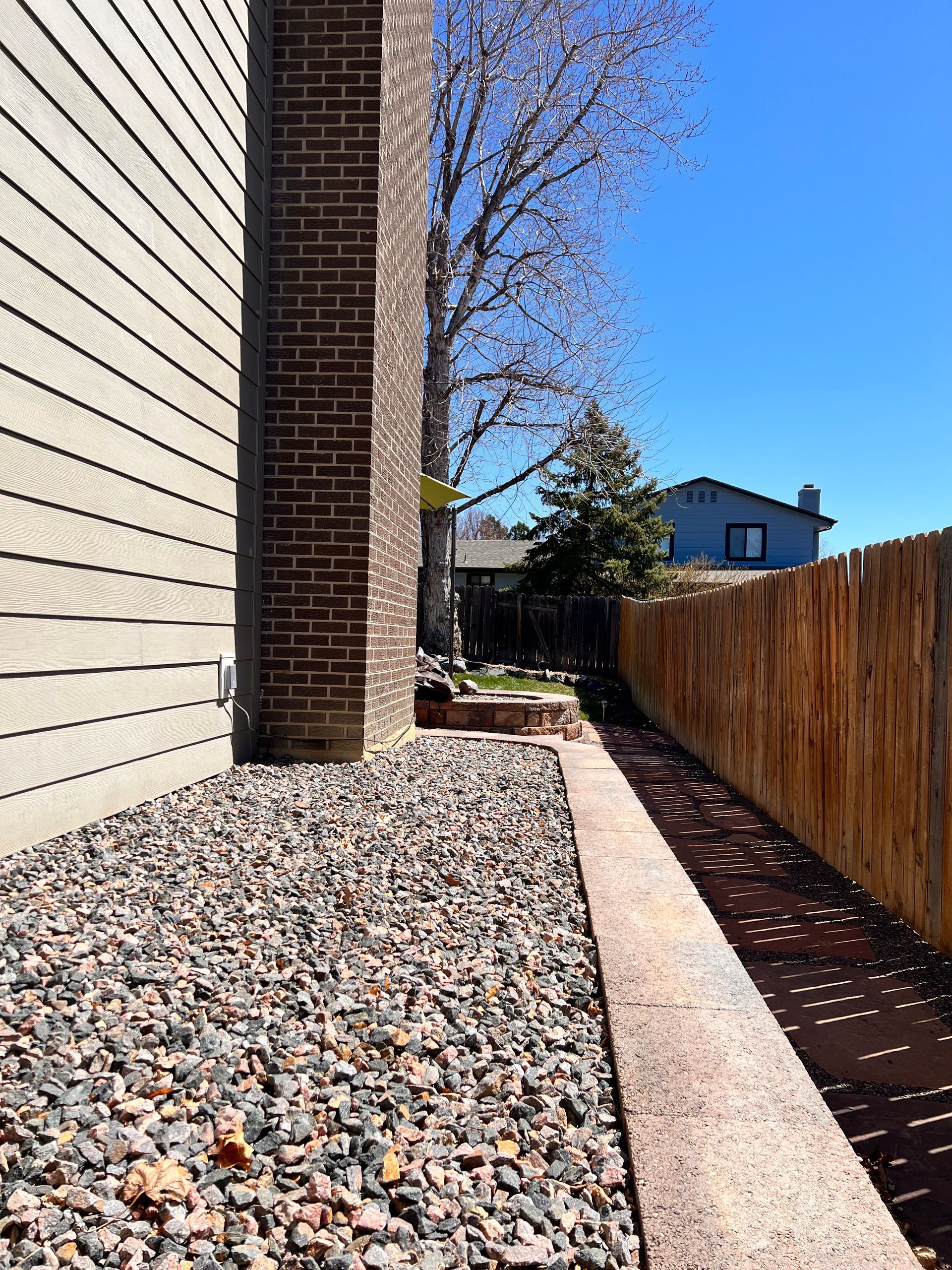 Exterior view: Brick chimney, gravel bed, wooden fence, narrow walkway, and a bright blue sky.