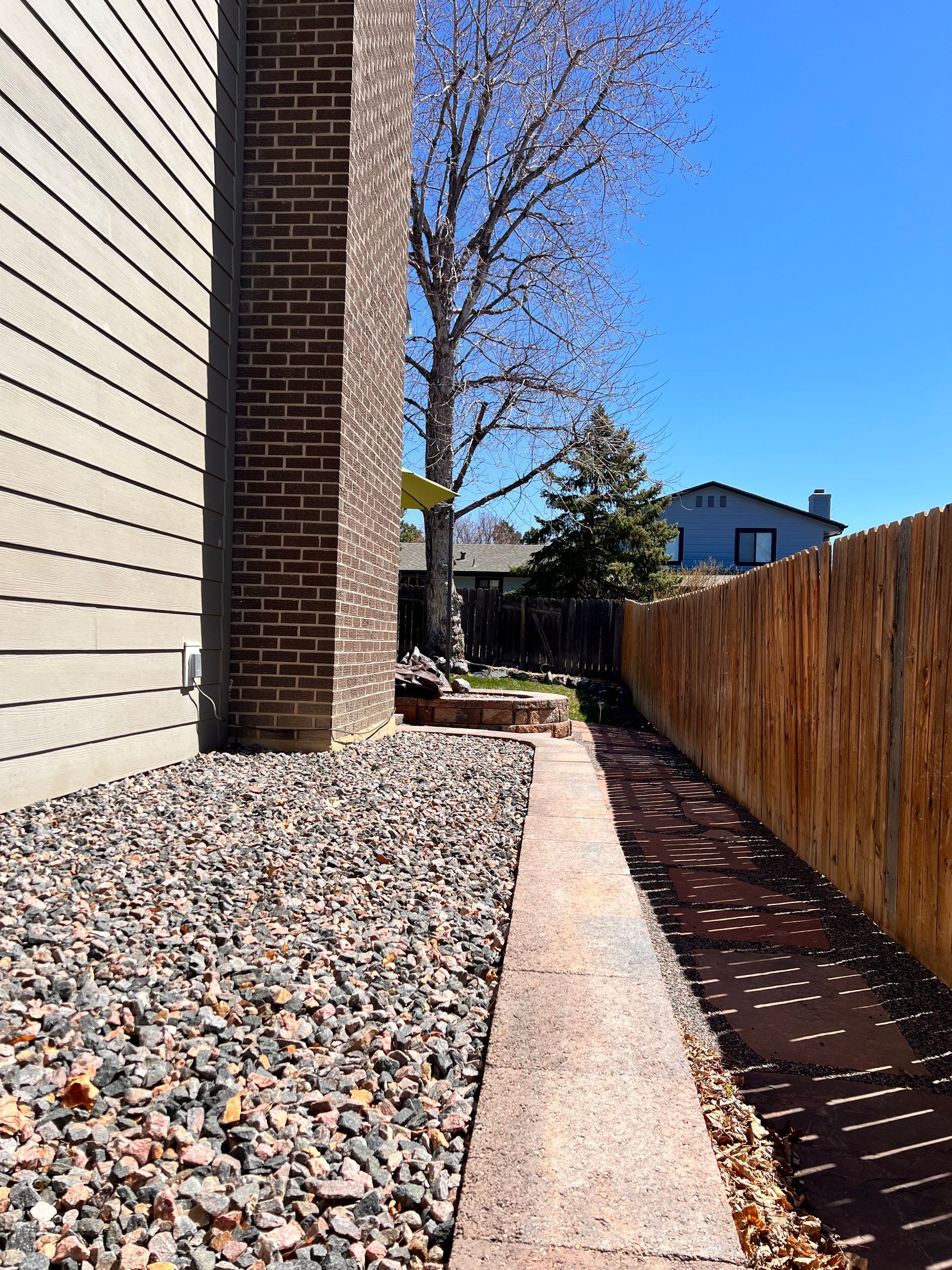 Narrow walkway alongside a building and fence, with gravel, pathway, and blue sky.