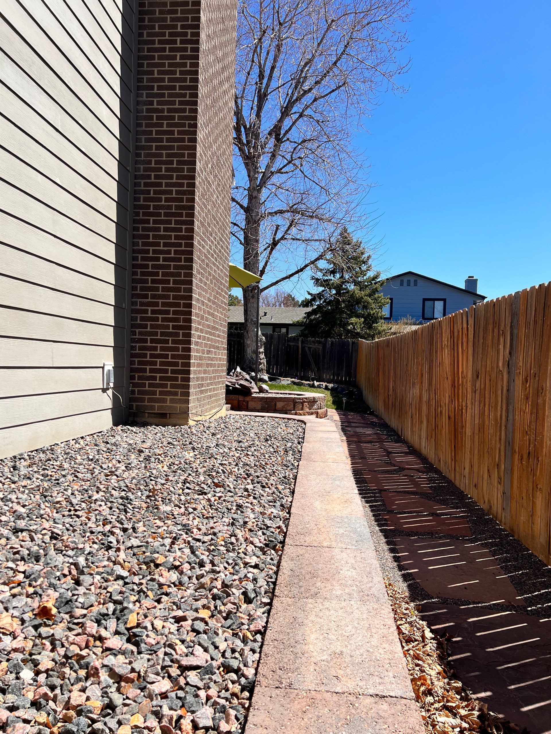 Narrow brick path between building, rock bed, and wooden fence on a sunny day.