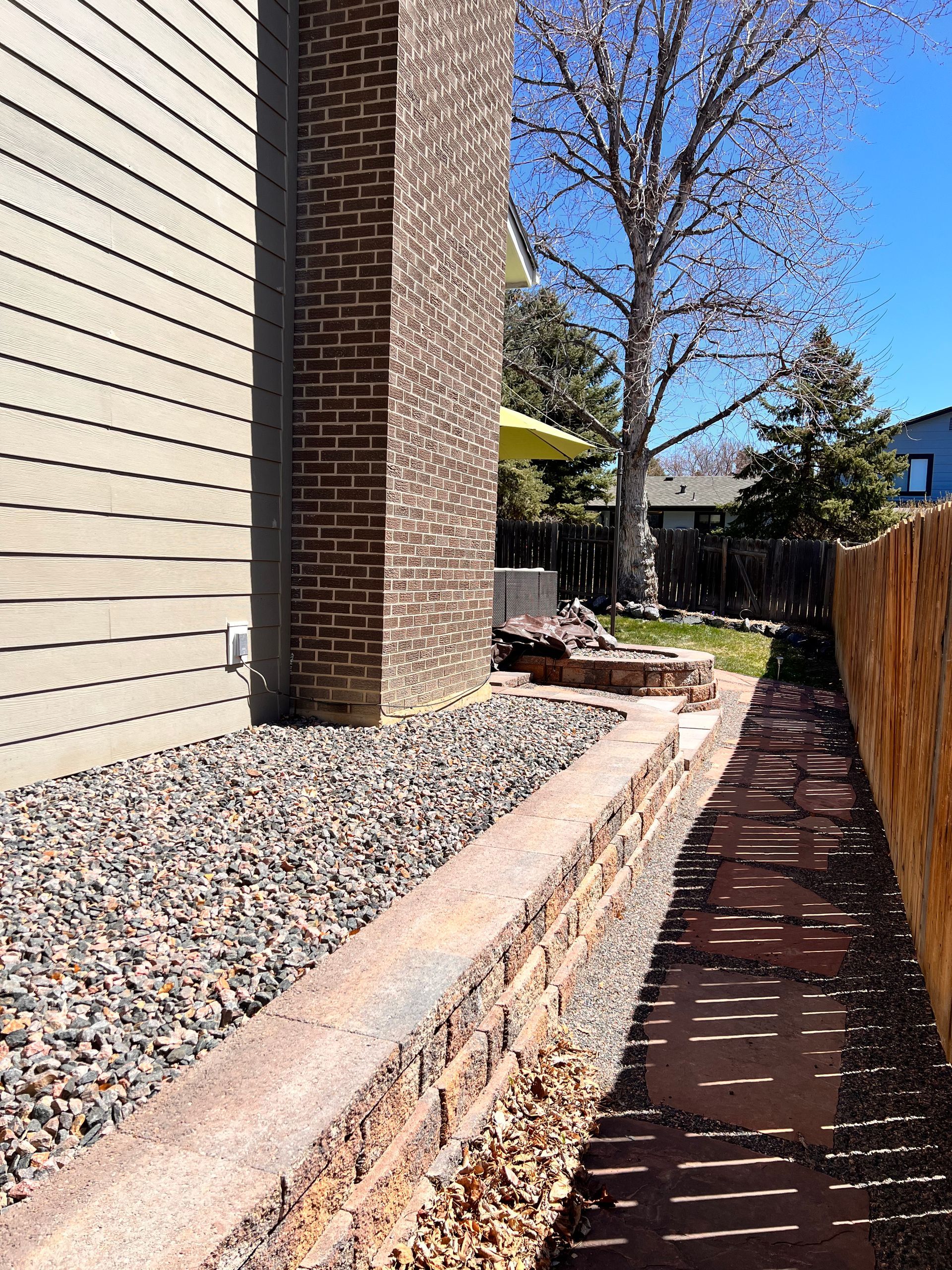 Exterior view: brick chimney next to a house with gravel bed, brick retaining wall, and narrow brick pathway alongside a wooden fence.