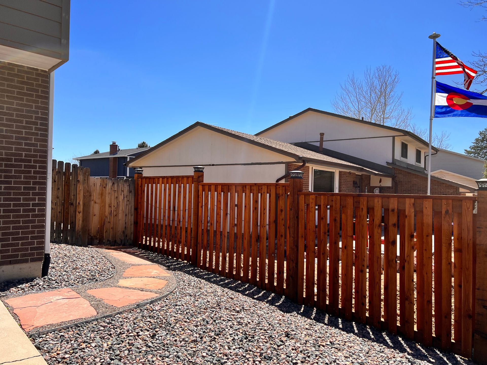 A fenced backyard with a flag, stone pathway, and gravel. Sunny day, blue sky.