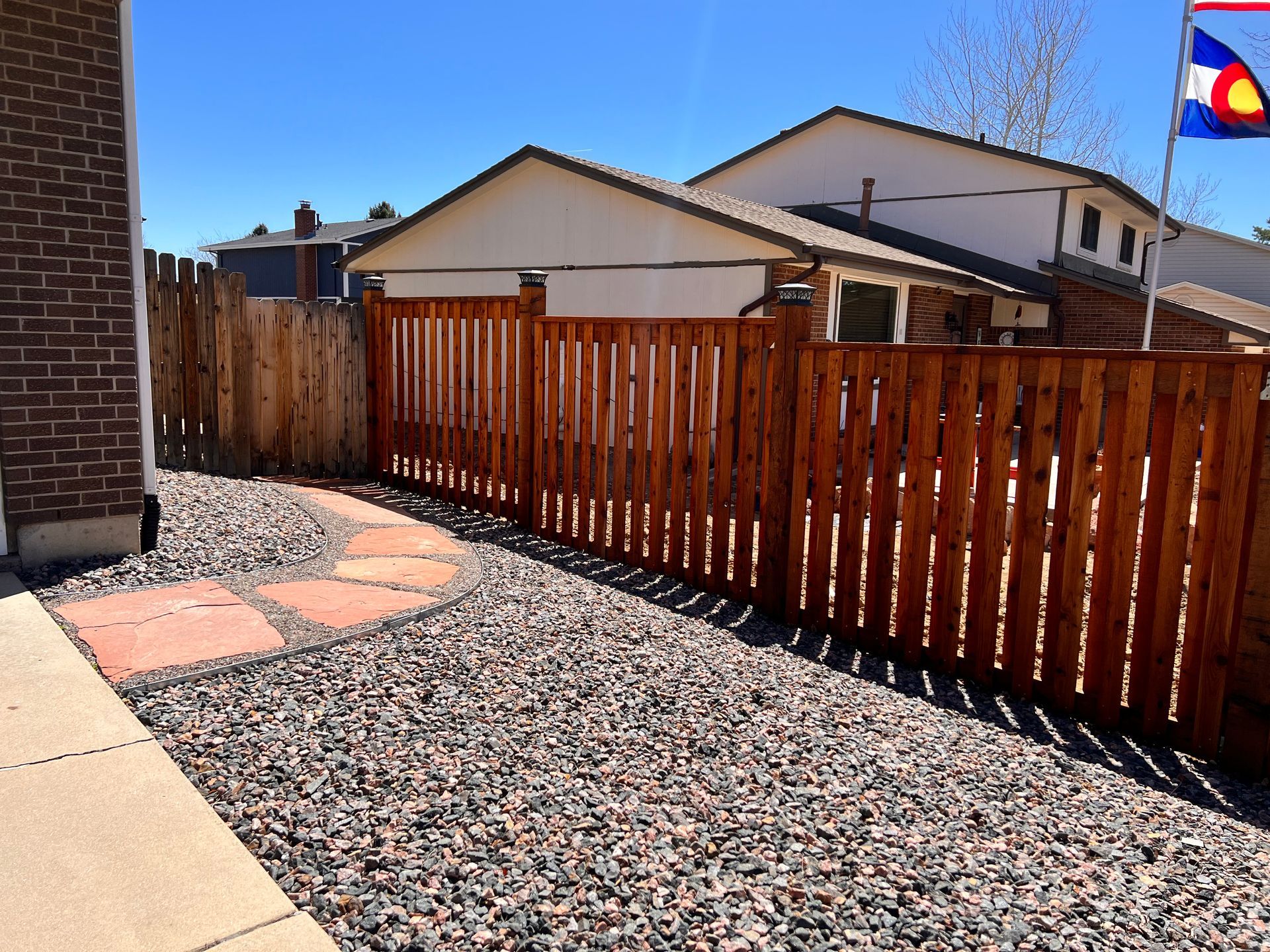 Backyard with reddish-brown fence, gravel, stepping stones, and Colorado flag on a sunny day.