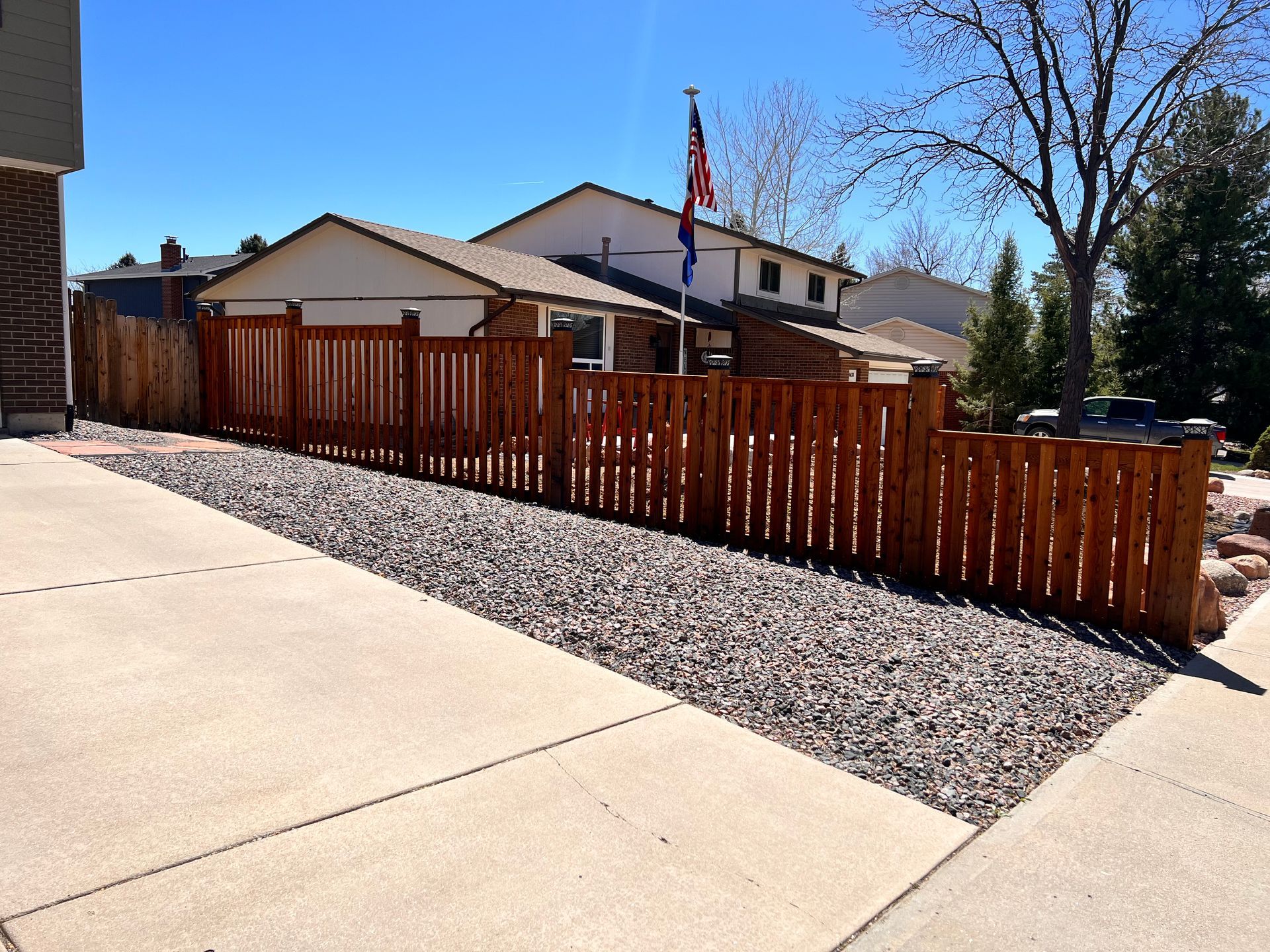 Brown wooden fence lines a driveway with gravel, houses, and an American flag on a sunny day.