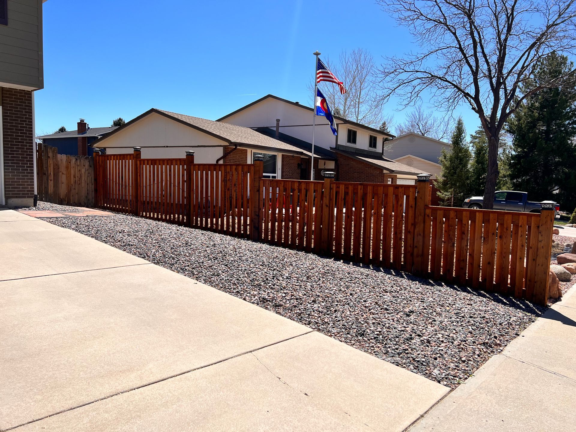 Wooden fence borders a driveway filled with gravel, a house with flags in the background.