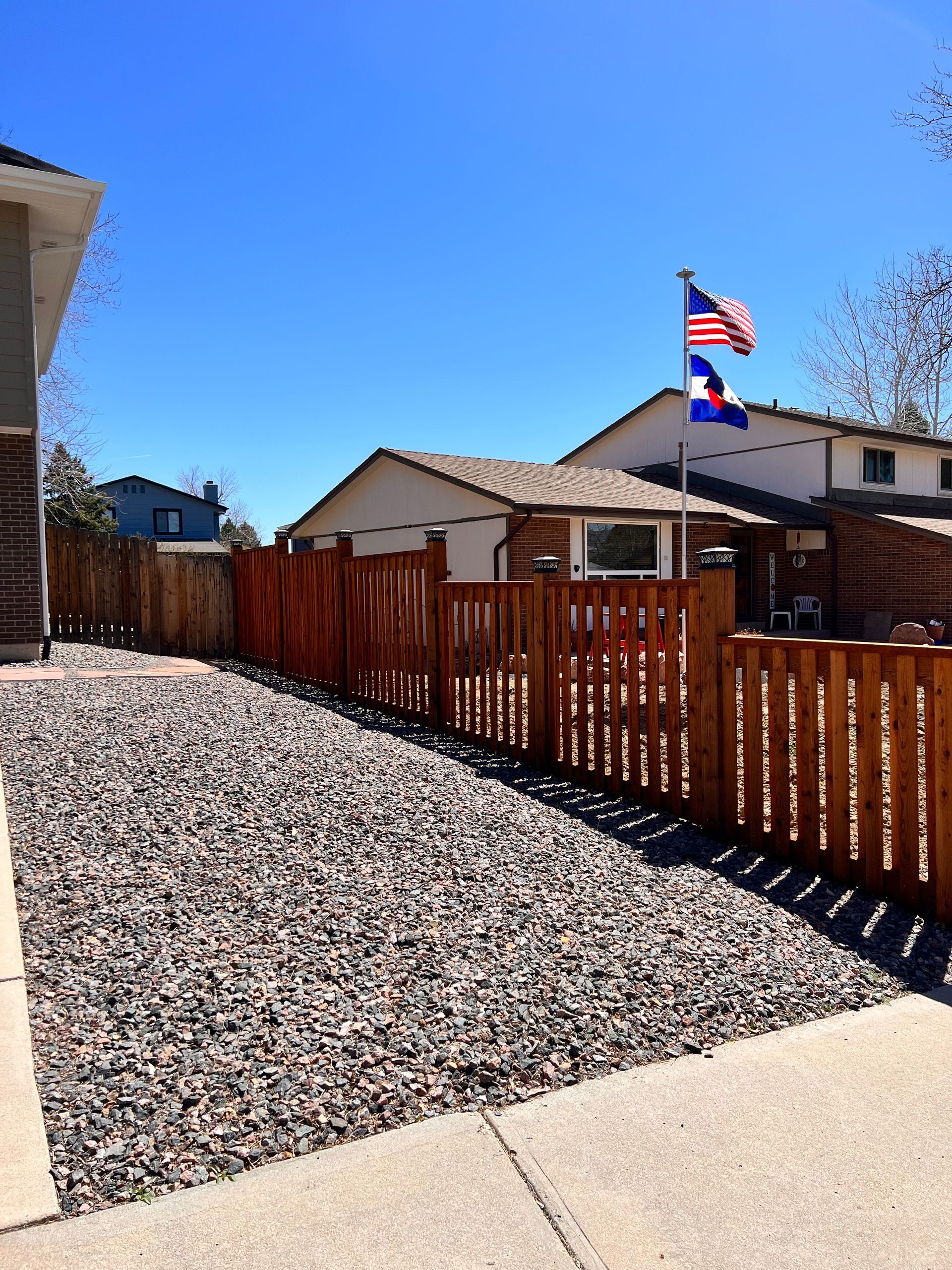 A wooden fence borders a gravel yard next to a sidewalk and a house under a clear, blue sky.