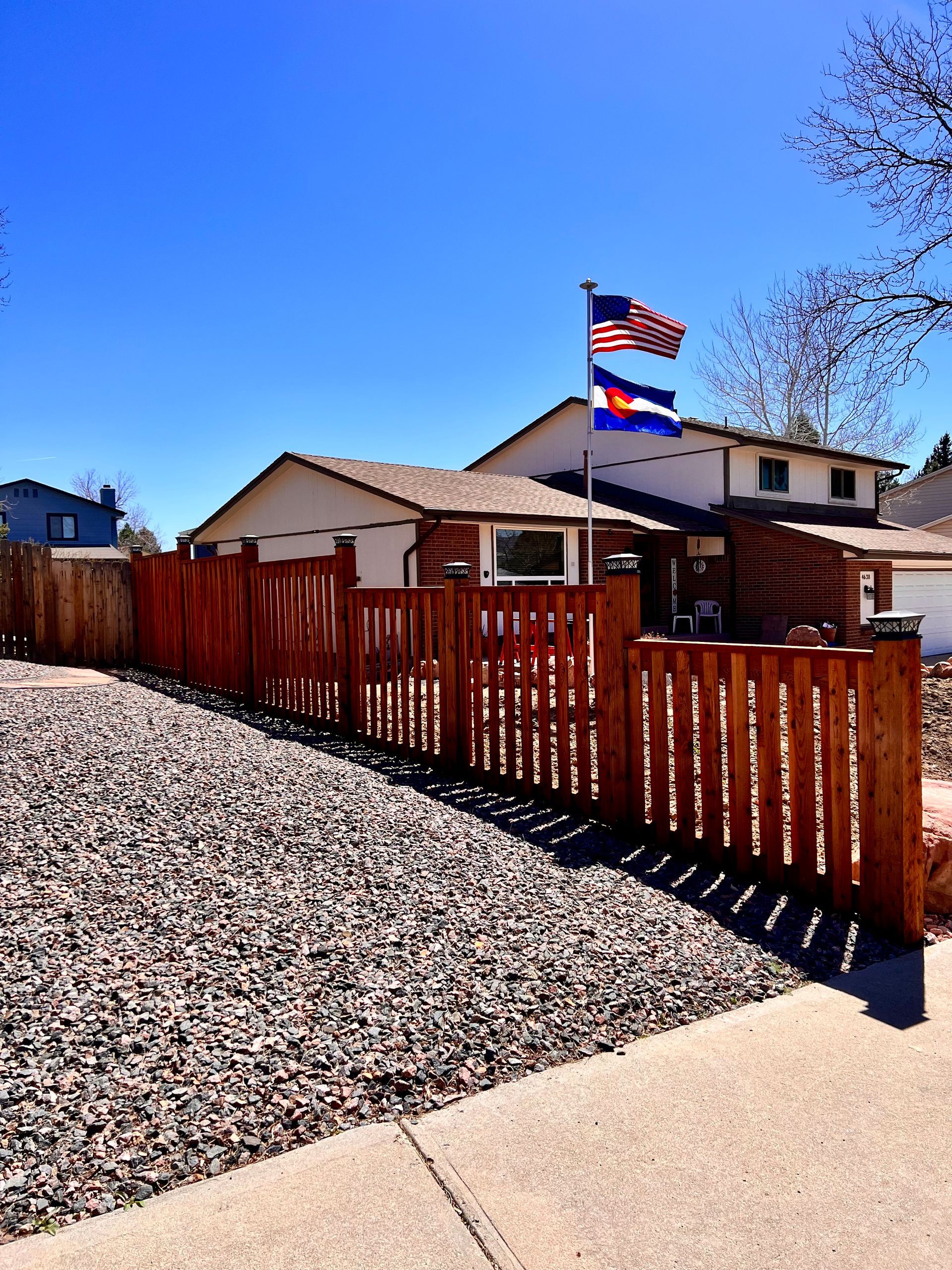 House with wooden fence, gravel driveway, and flags of the USA and Colorado under a bright blue sky.
