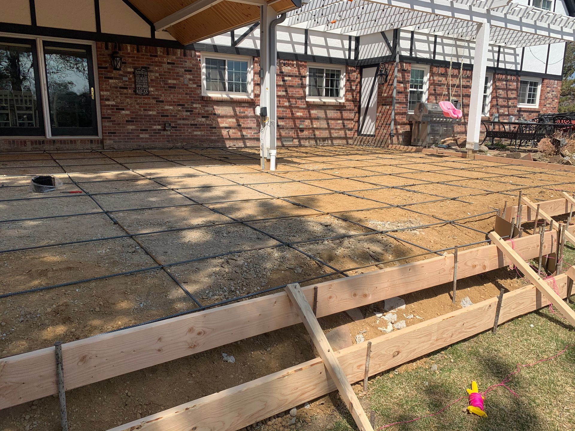 Construction site: wooden forms, gravel, and rebar for a patio, in front of a brick house.