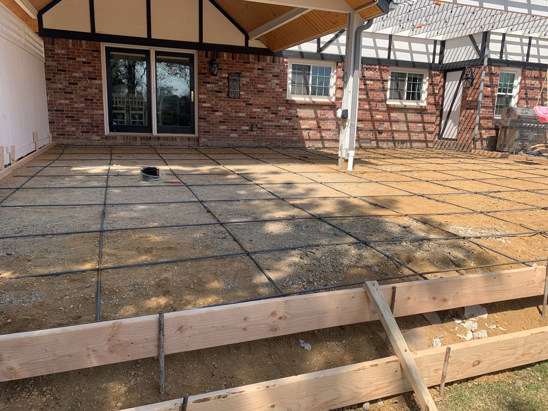 Concrete patio under construction with rebar grid and wooden formwork. Brick house background.