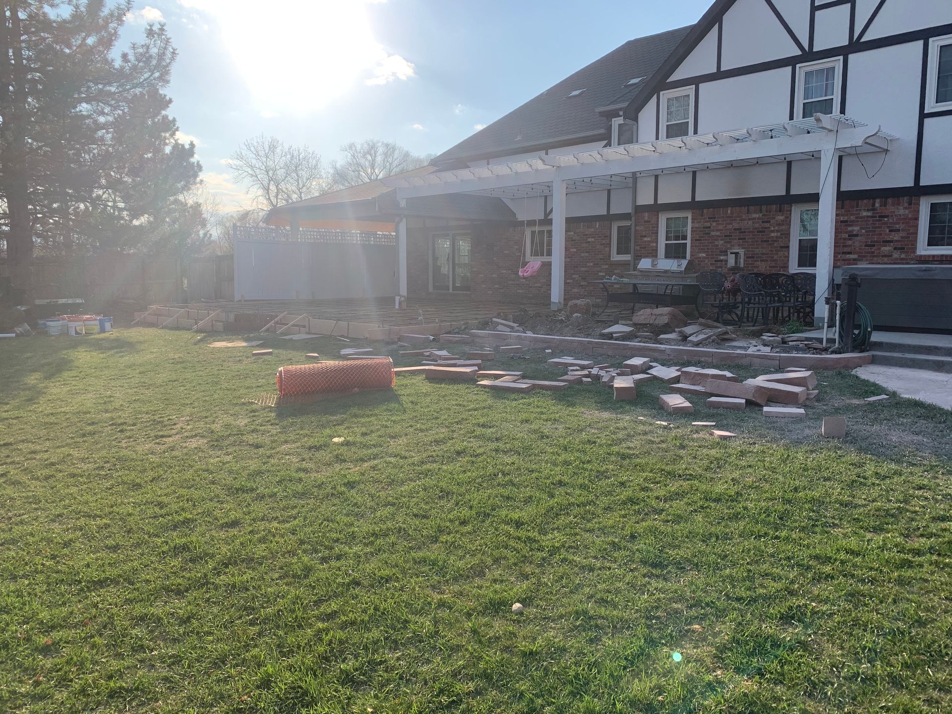 Backyard scene with house, pergola, scattered bricks, and green grass.
