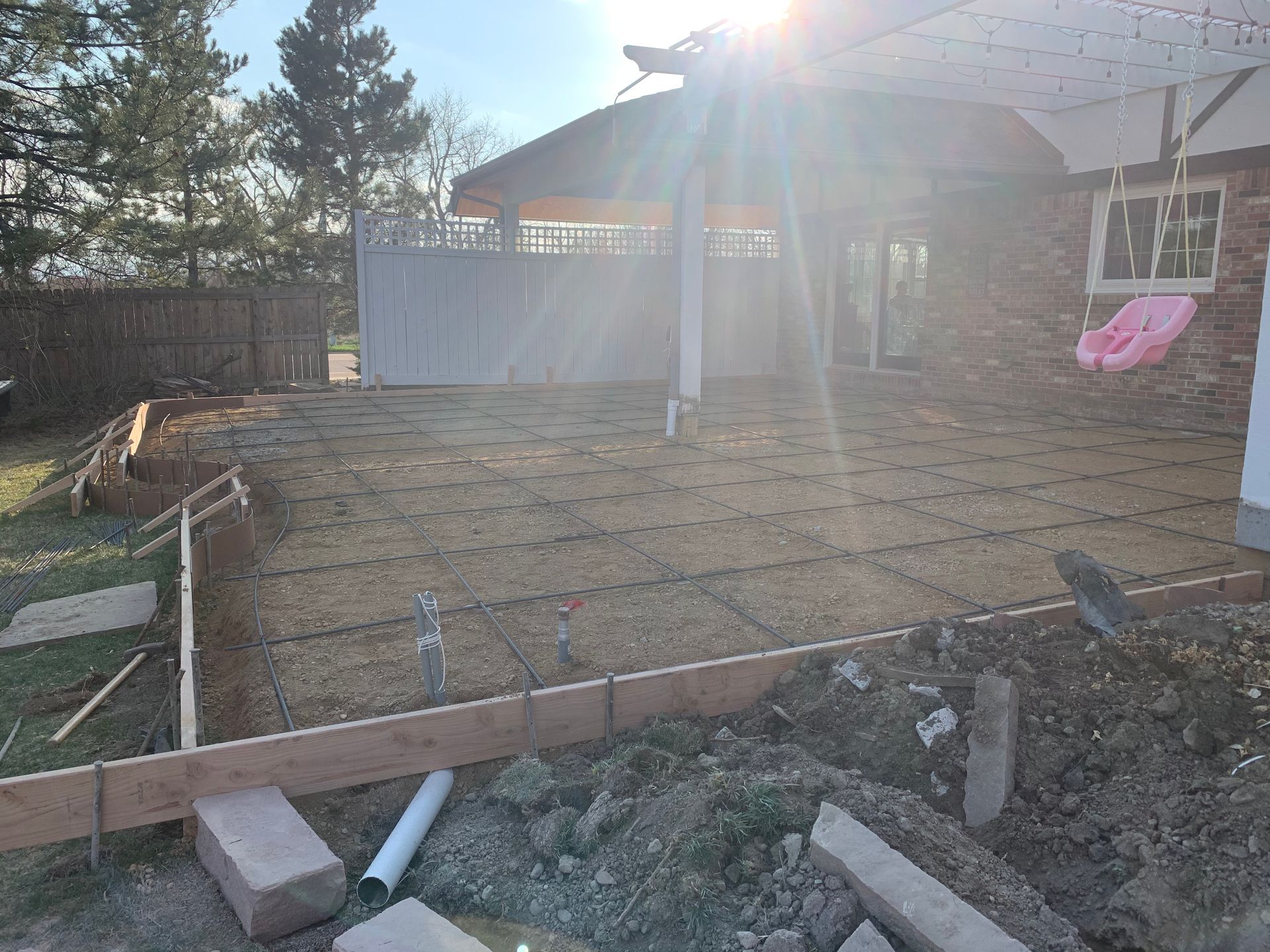 Construction site: concrete patio formwork, rebar grid, dirt pile, sun glare.
