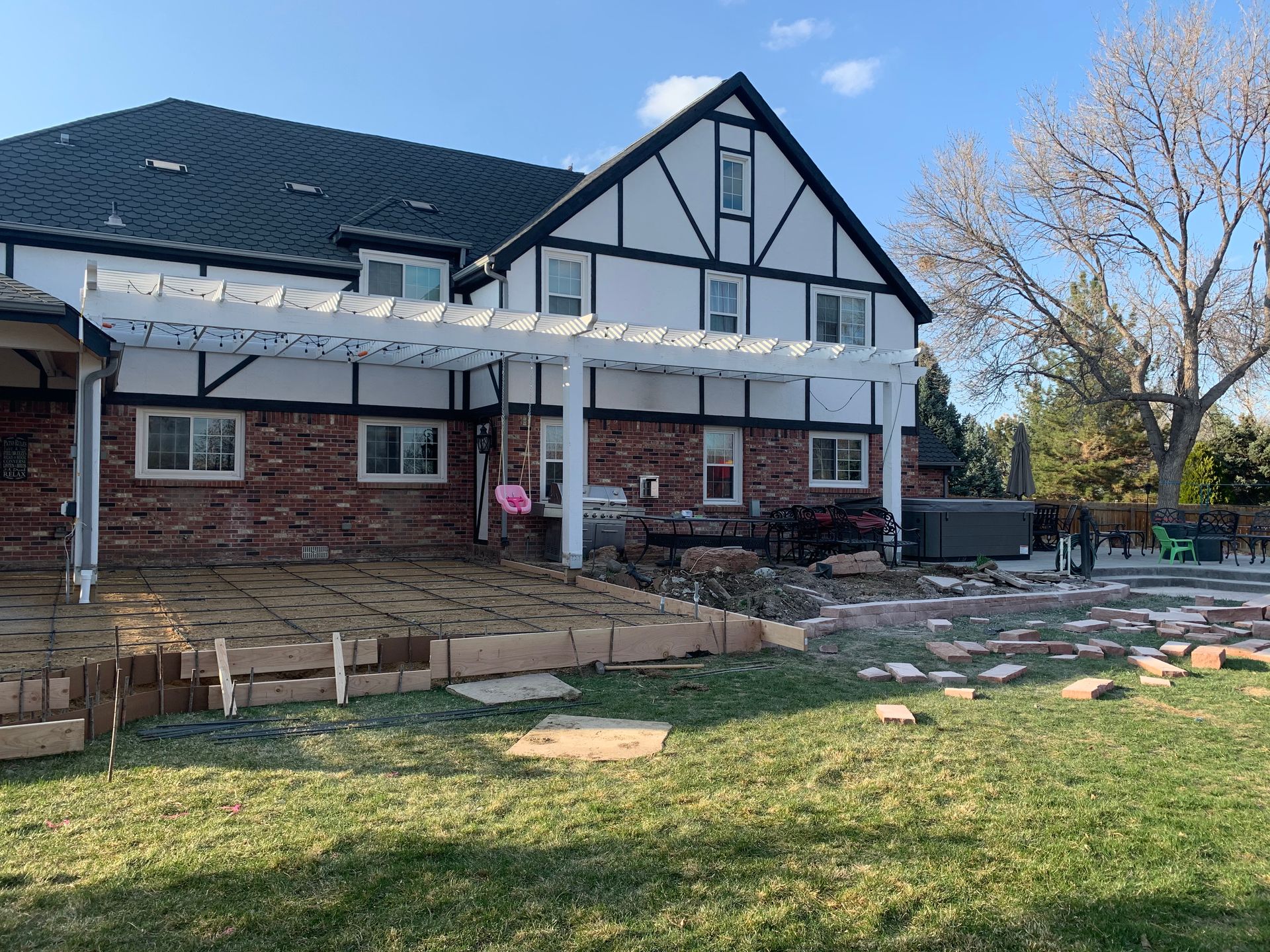 Backyard deck construction next to a Tudor-style brick house with pergola. Lawn in the foreground.