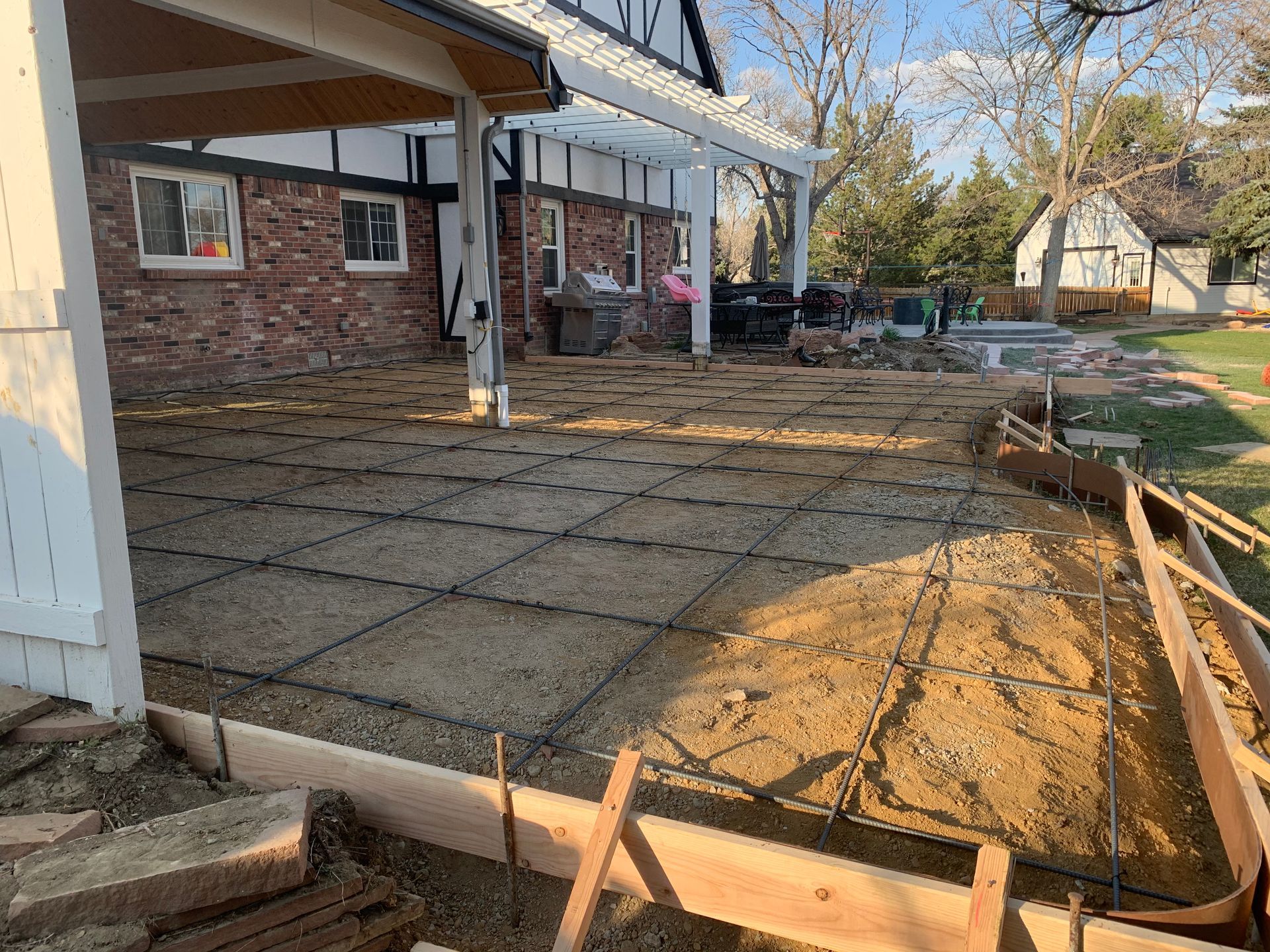 Construction site: Concrete patio being prepared with rebar grid; house in background.