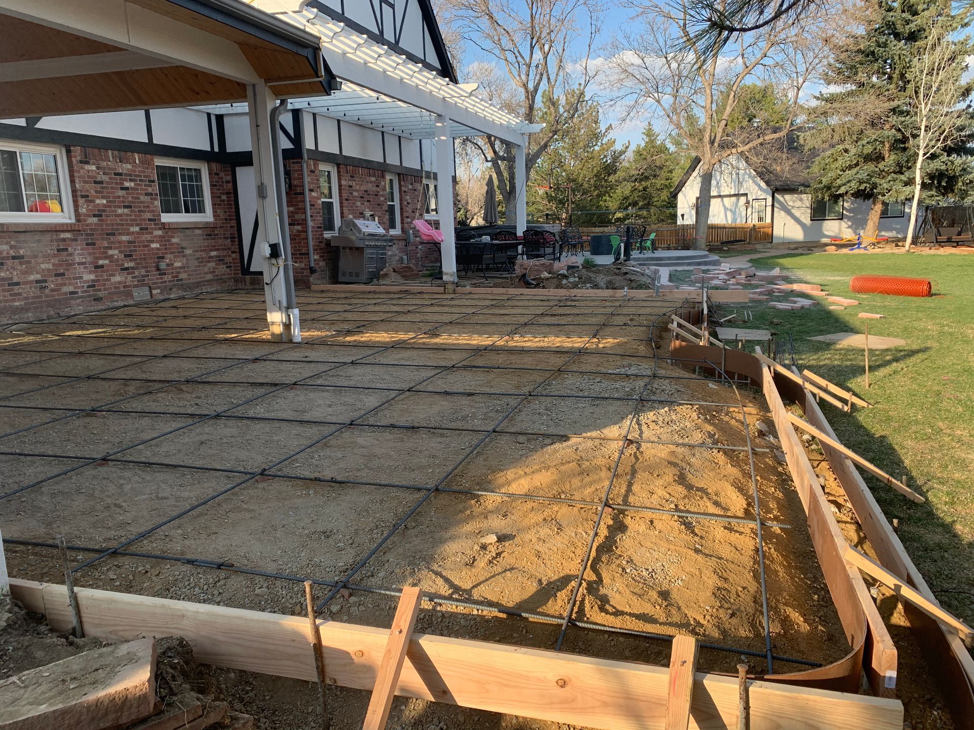 Construction site: concrete patio with rebar grid, wooden forms, and a house in the background.