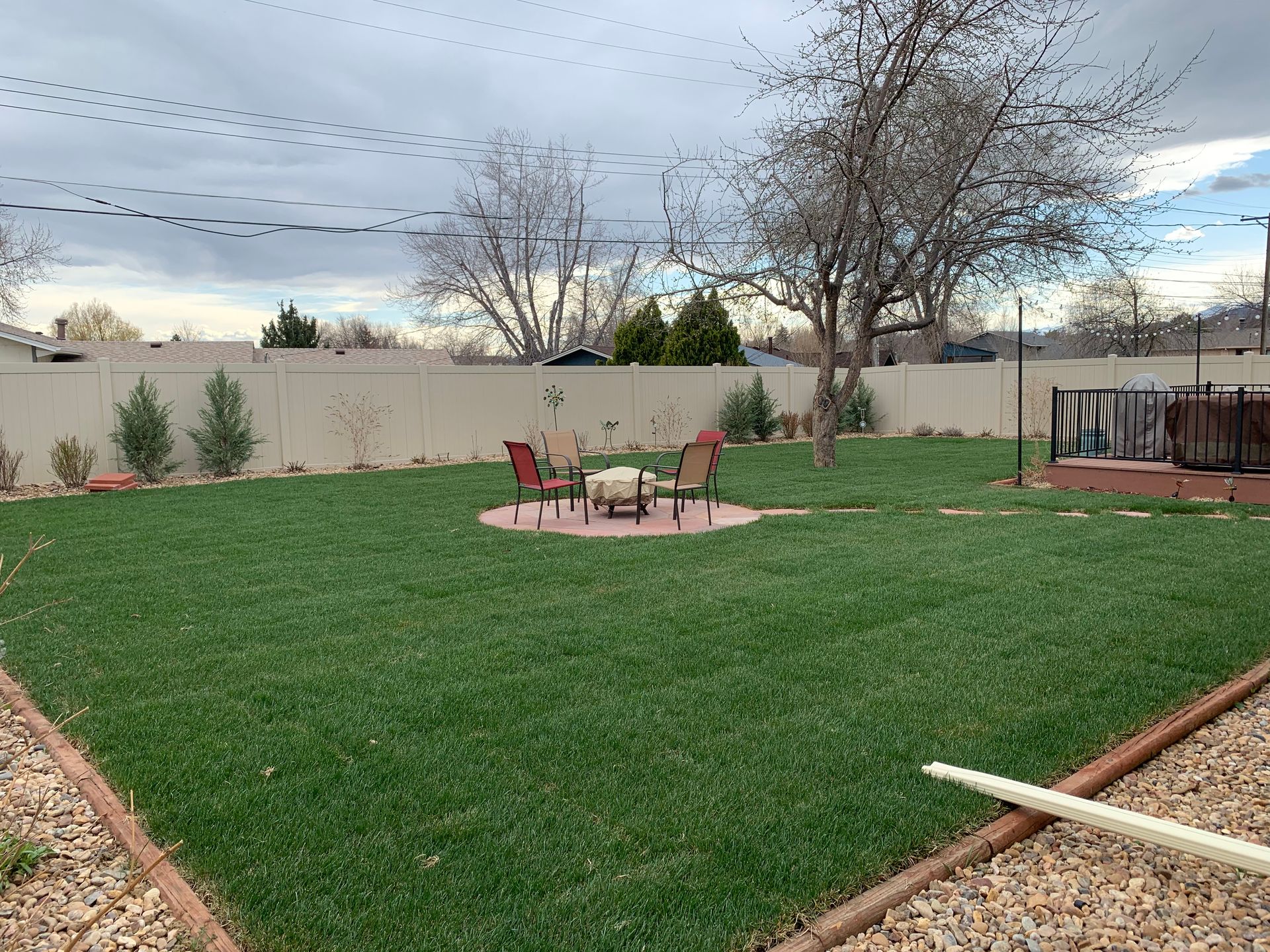 Well-manicured backyard with green grass, seating around a fire pit, and a white fence.