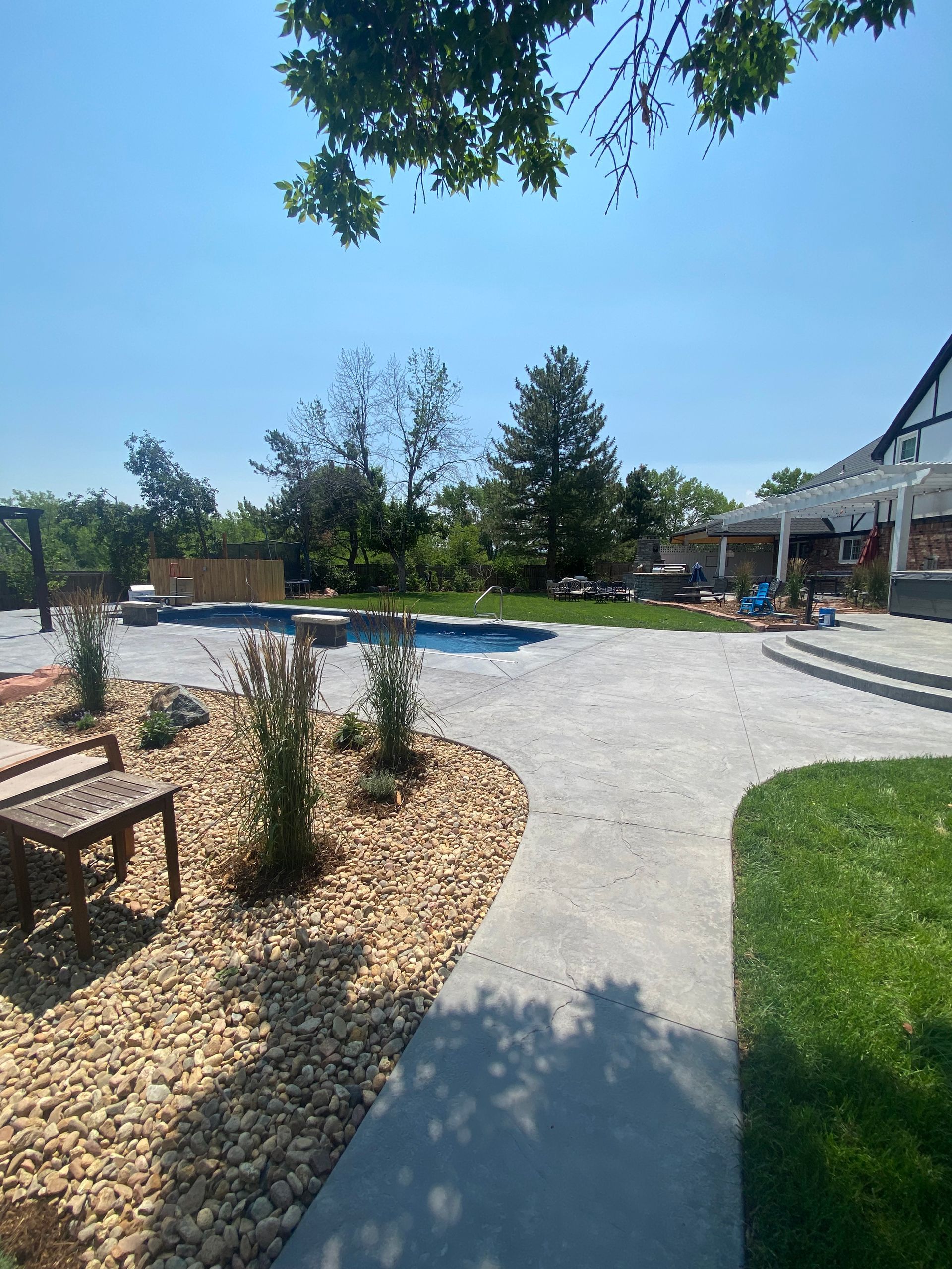 Backyard with pool, patio, landscaping, and a house in the background on a sunny day.
